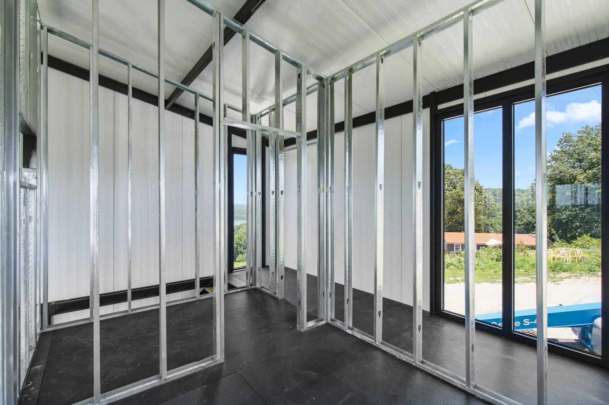 Interior of a house under construction with metal framing, large windows showing trees and houses outside, and a black floor.