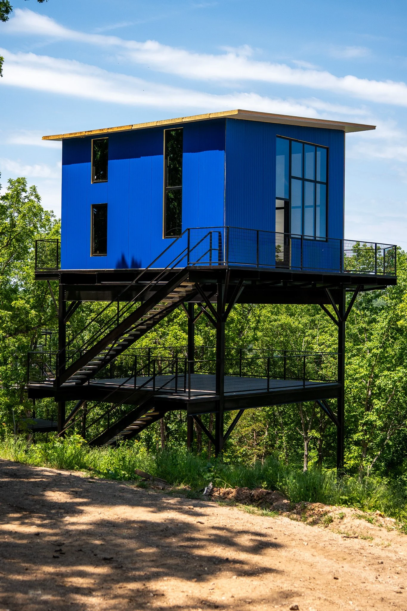 A modern, elevated house with a blue exterior and large glass windows, surrounded by trees and greenery under a blue sky with clouds.