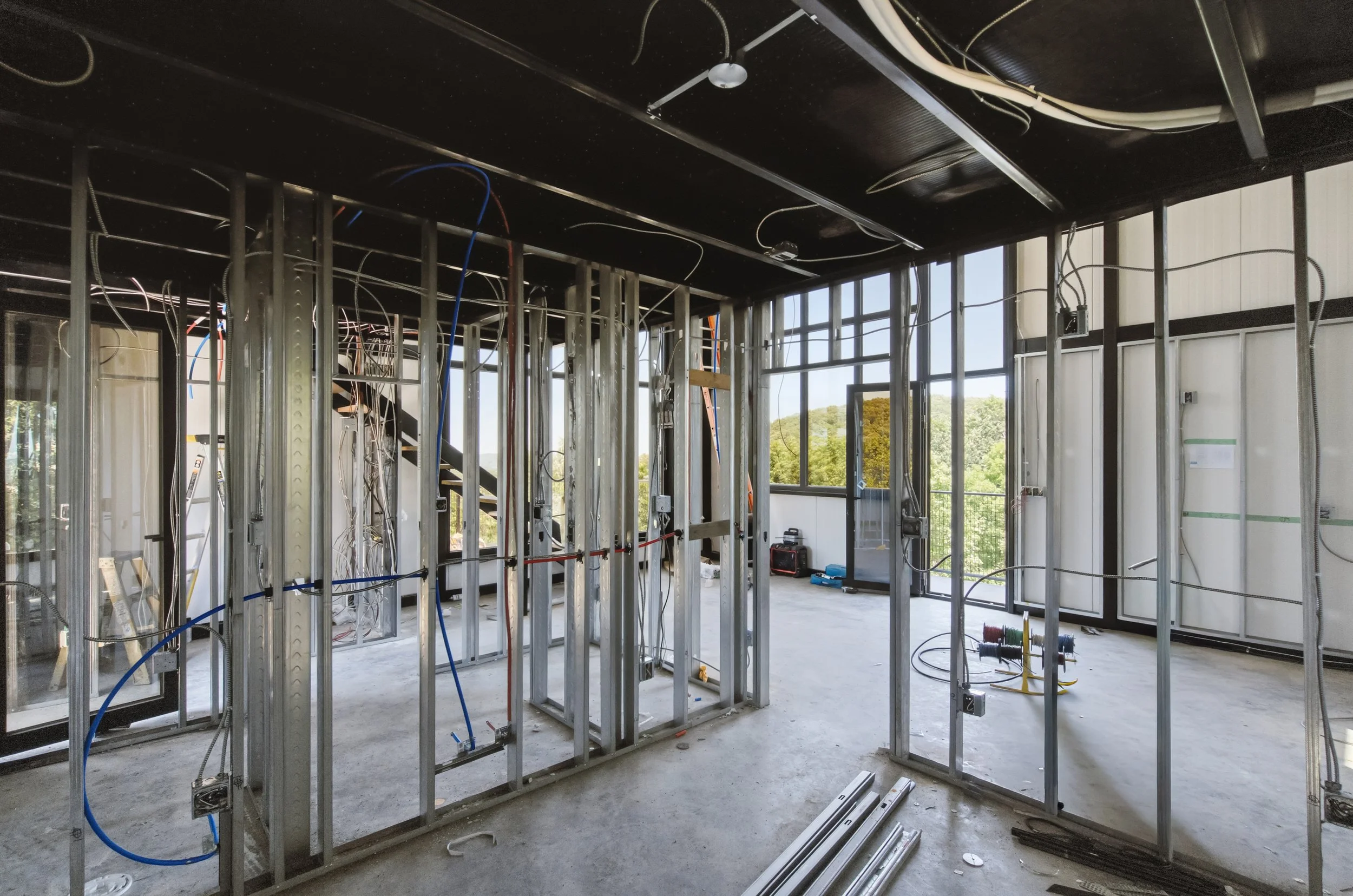Interior framing of a building under construction with exposed metal studs, electrical wiring, and large windows showing trees outside.
