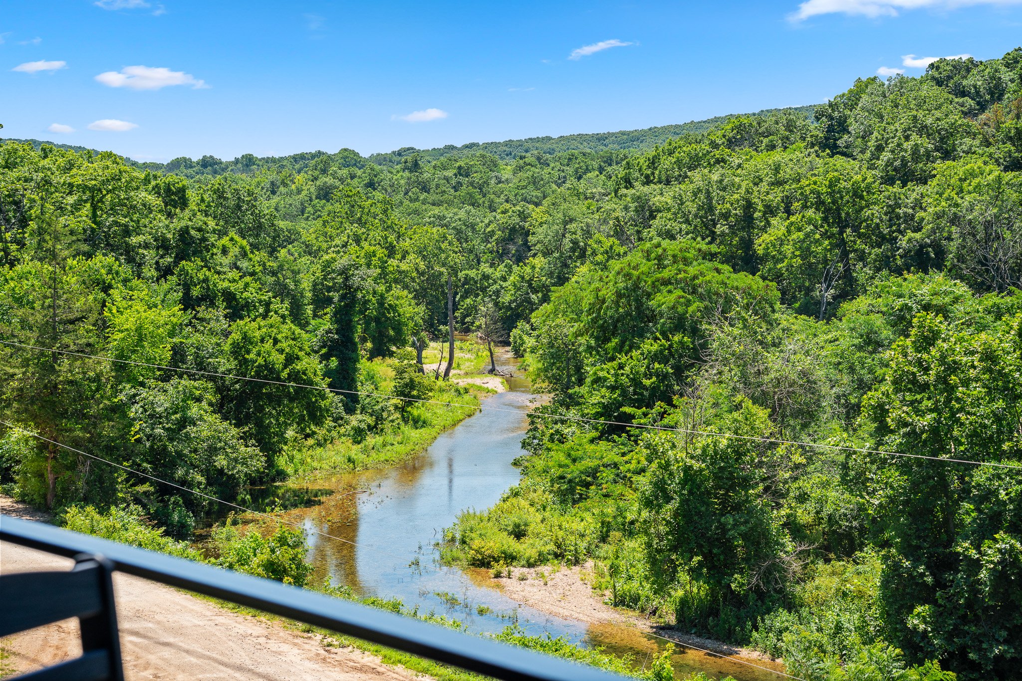 A river flowing through a lush, green forest with hills in the background under a blue sky with a few clouds.