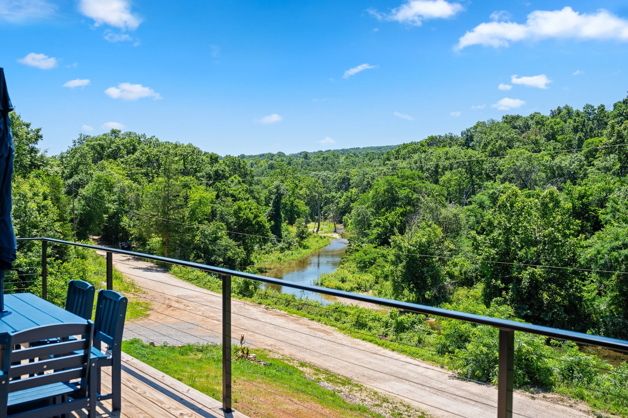 A scenic landscape Viewed from a porch or balcony, overlooking a forested area with a small winding river below, under a blue sky with scattered clouds.
