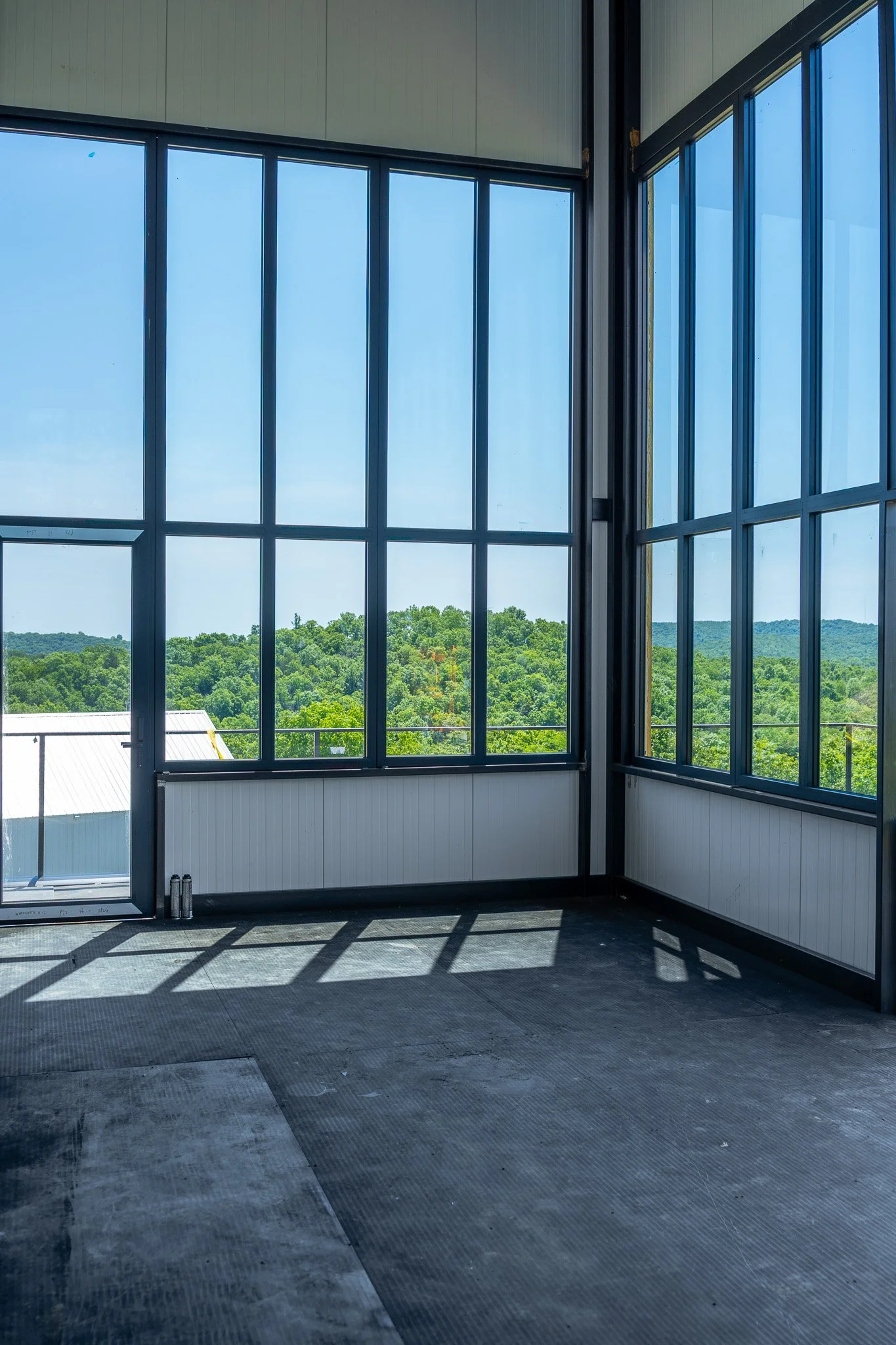 Interior view of a building with large glass windows showing a green landscape outside, and shadows cast on the black rubber flooring inside.