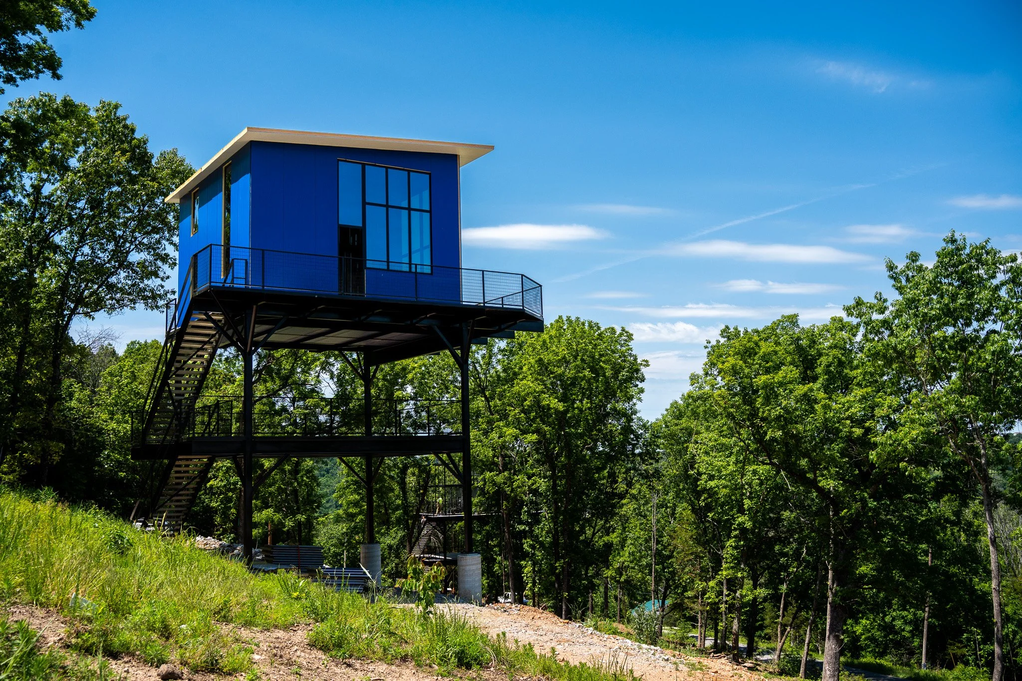 A modern blue elevated house on stilts in a wooded area with green trees and a clear blue sky.