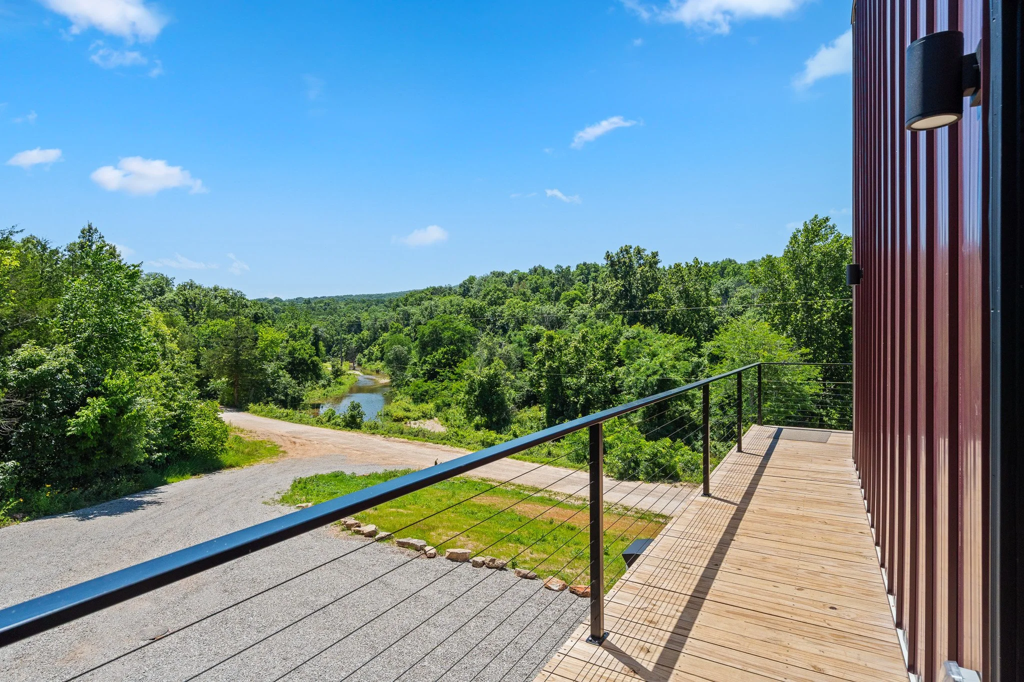 View from a building's balcony overlooking a rural landscape with a dirt road, green trees, a small river, and a blue sky with scattered clouds.