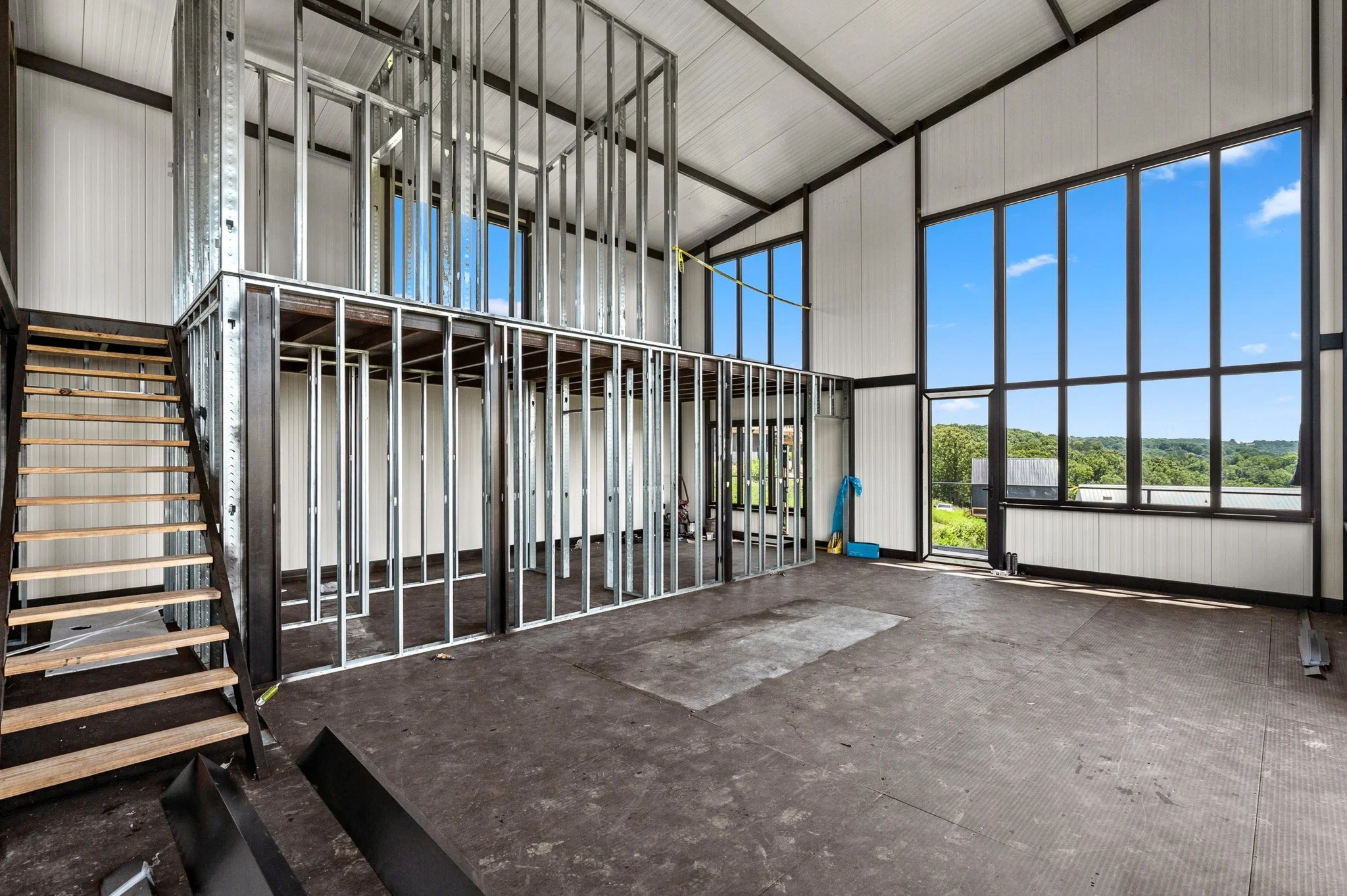Interior of a house construction site with metal framing for walls and stairs, large windows showing a view of trees and blue sky.