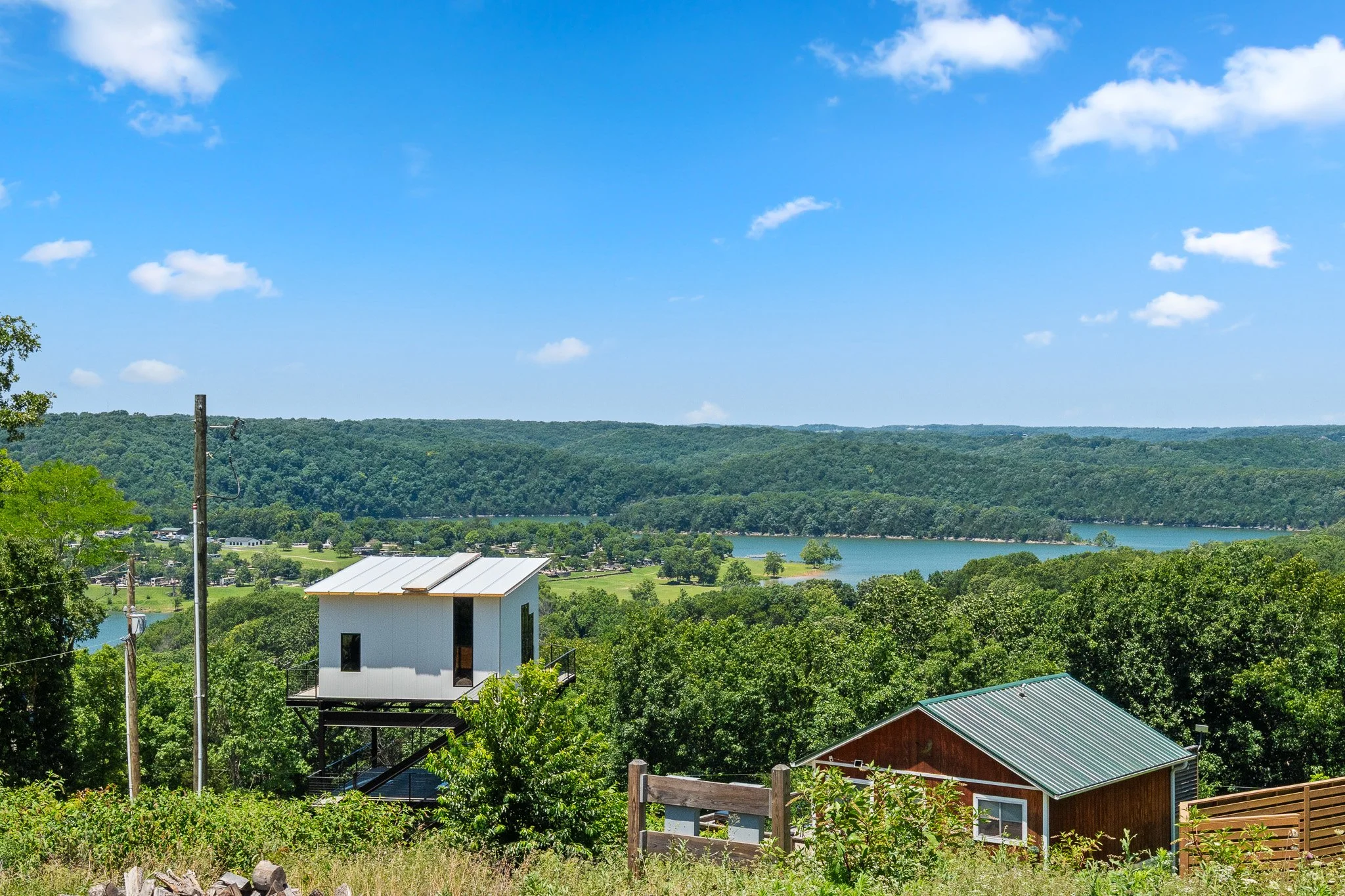 A scenic view of a lake with a backdrop of green hills under a partly cloudy blue sky. In the foreground, there's a white building on stilts and other houses with metal roofs surrounded by trees and greenery.