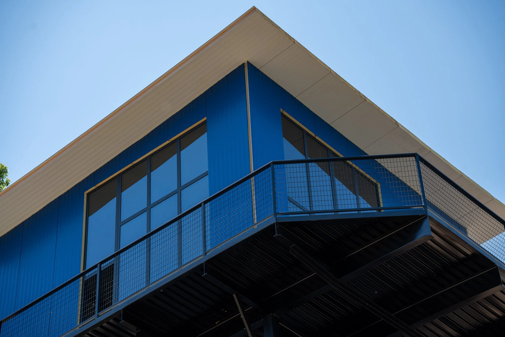 Close-up of a modern building's corner with blue exterior walls, large glass windows, black metal railing, and a light-colored overhanging roof against a clear blue sky.