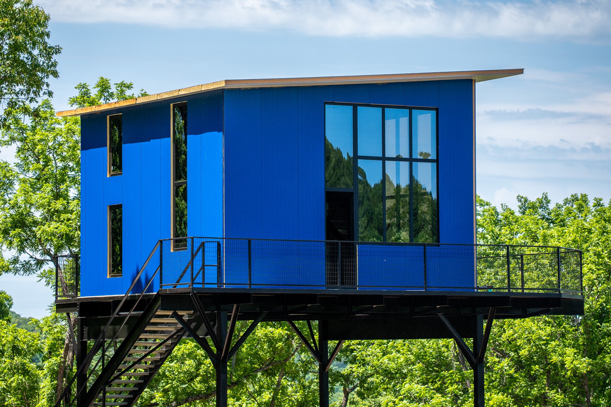 Modern blue house with large windows, set among green trees, with a metal staircase leading up to a balcony
