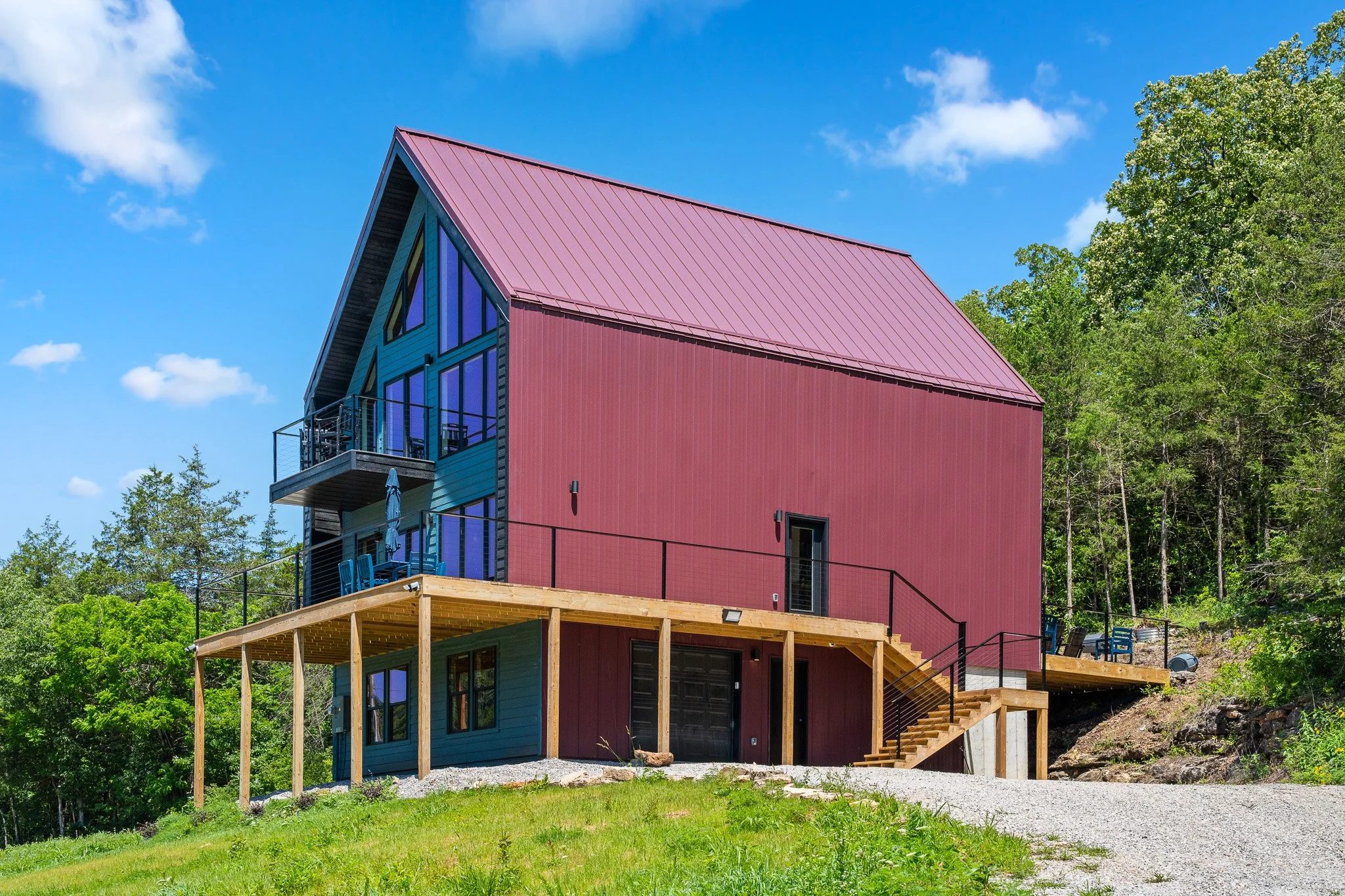 A modern house with blue siding and a red metal roof, built on a hillside with a deck and outdoor seating, surrounded by green trees under a blue sky.