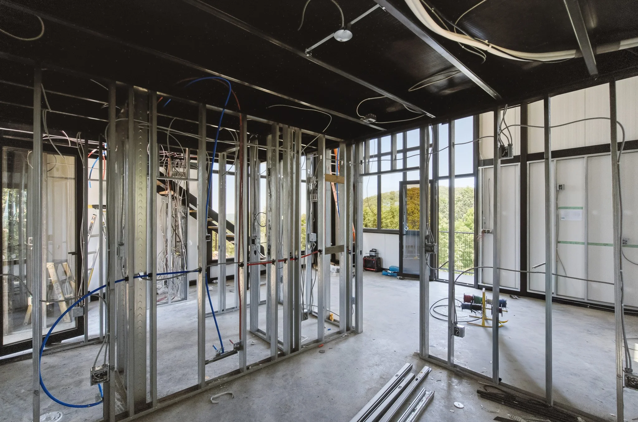Interior of a building under construction with exposed metal framing, electrical wiring, and large windows showing a view of trees outside.