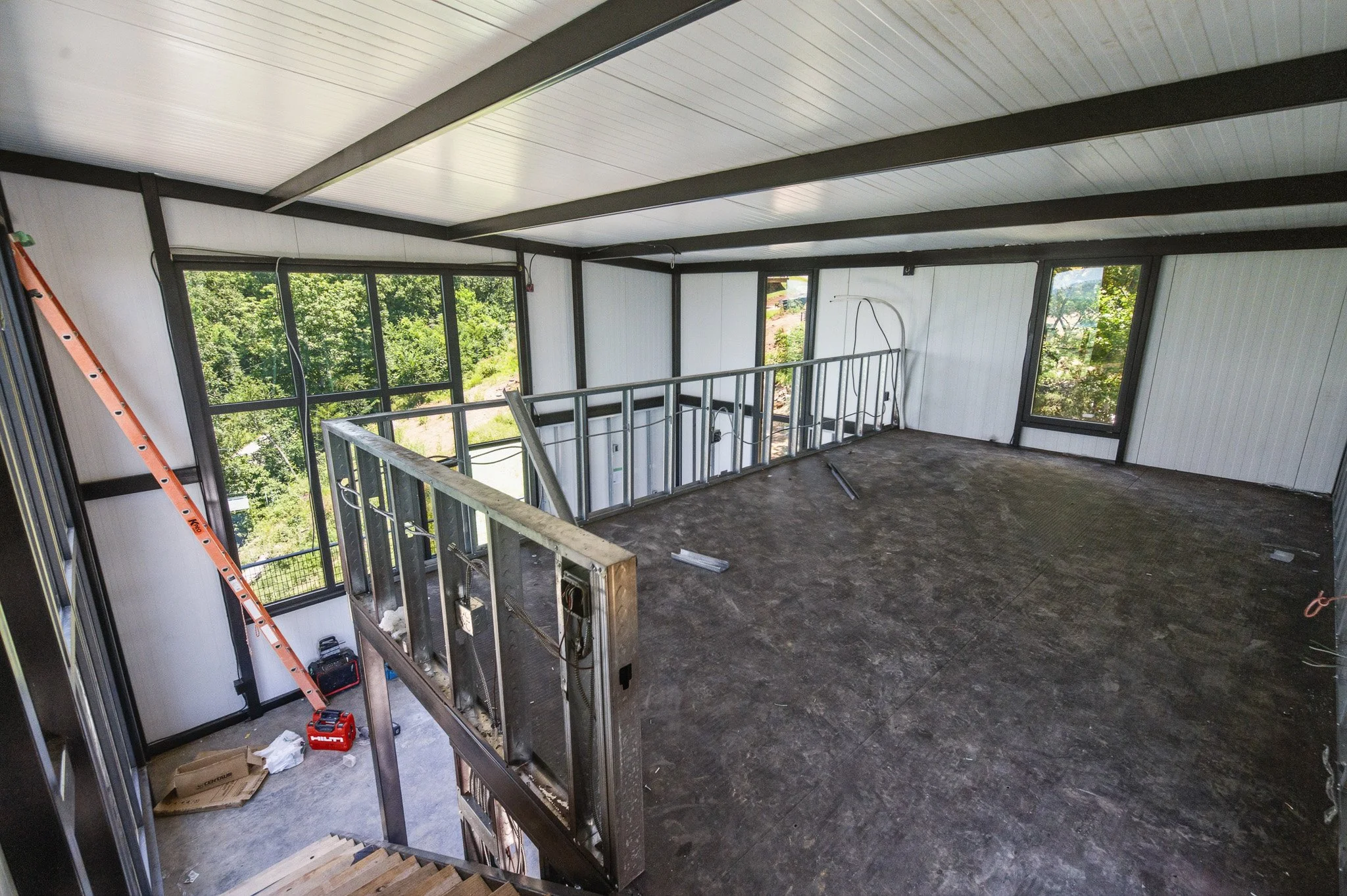 Interior view of a house under construction with exposed framing, a staircase, and large windows showing greenery outside.