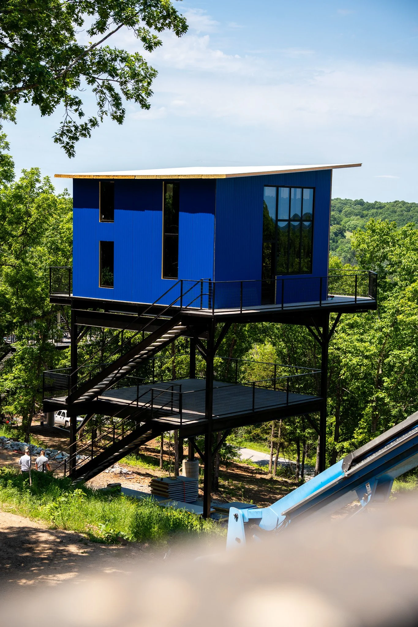 Under construction blue treehouse with large glass windows, timber roof, and black metal railings, surrounded by green trees and a clear blue sky.