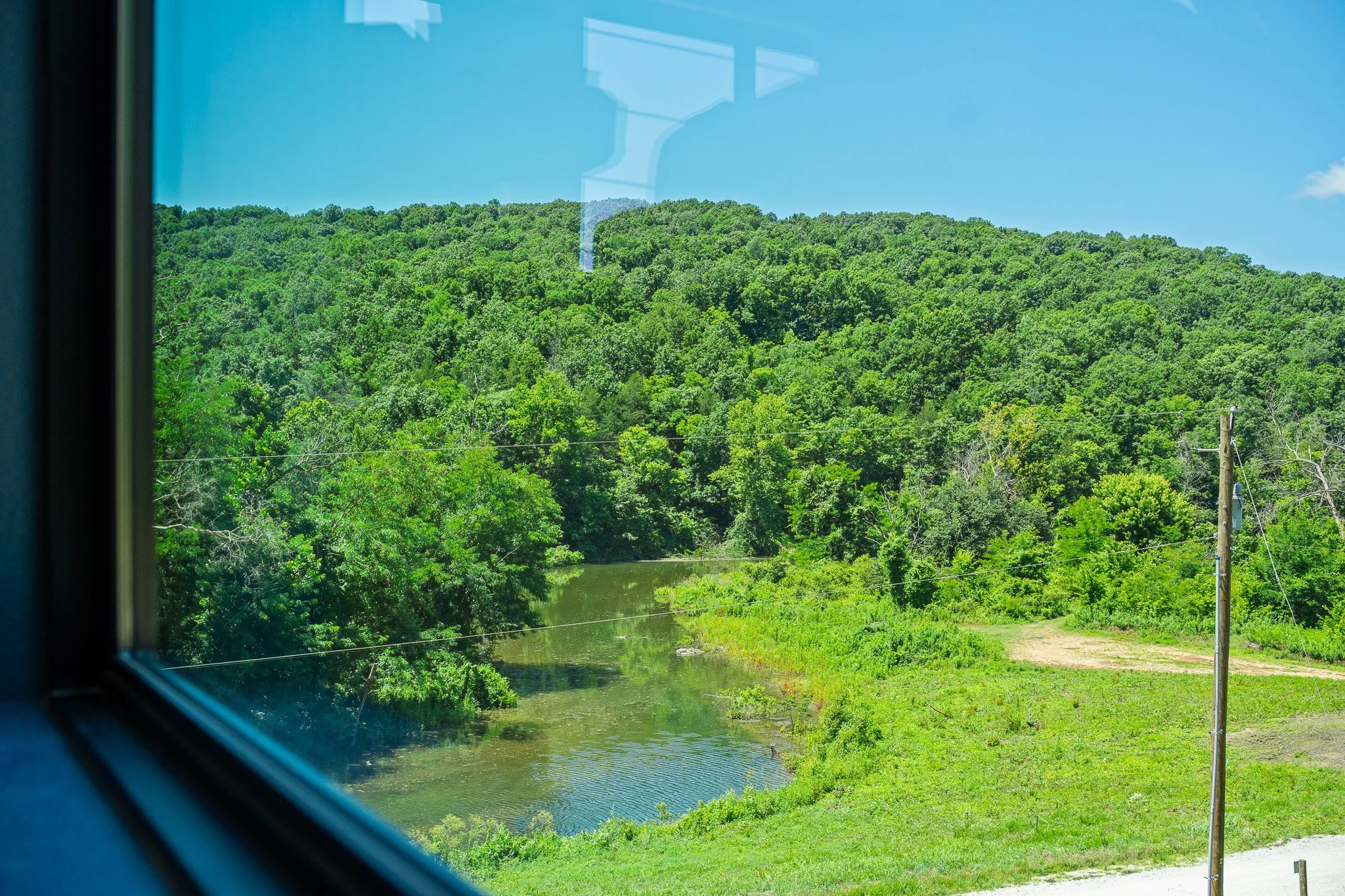 View through a window showing a lush green hillside, a small river or pond, and a clear blue sky with a few clouds. There are utility poles and power lines in the foreground.
