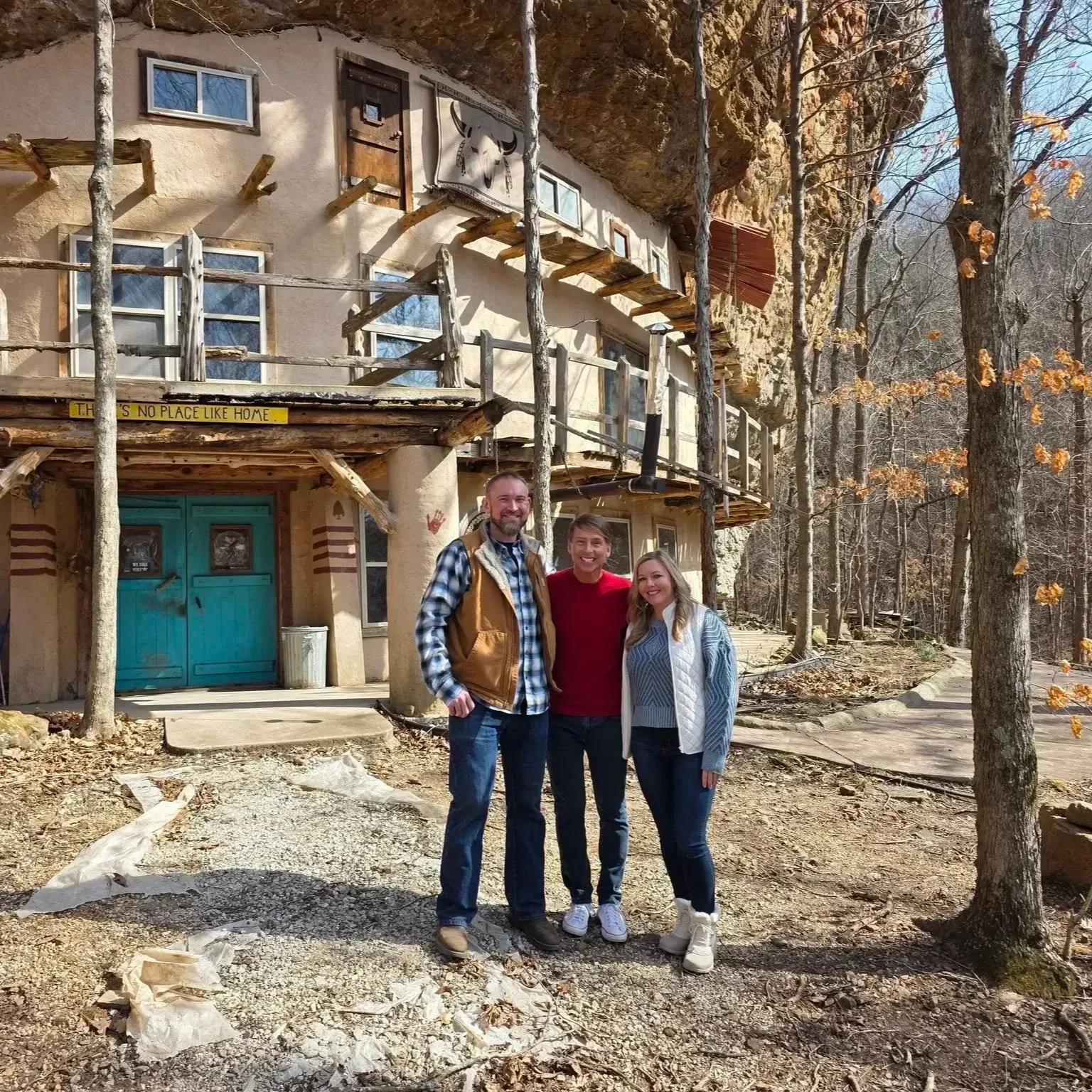 Three people stand in front of a rustic, house built into a cliffside with trees around, smiling.