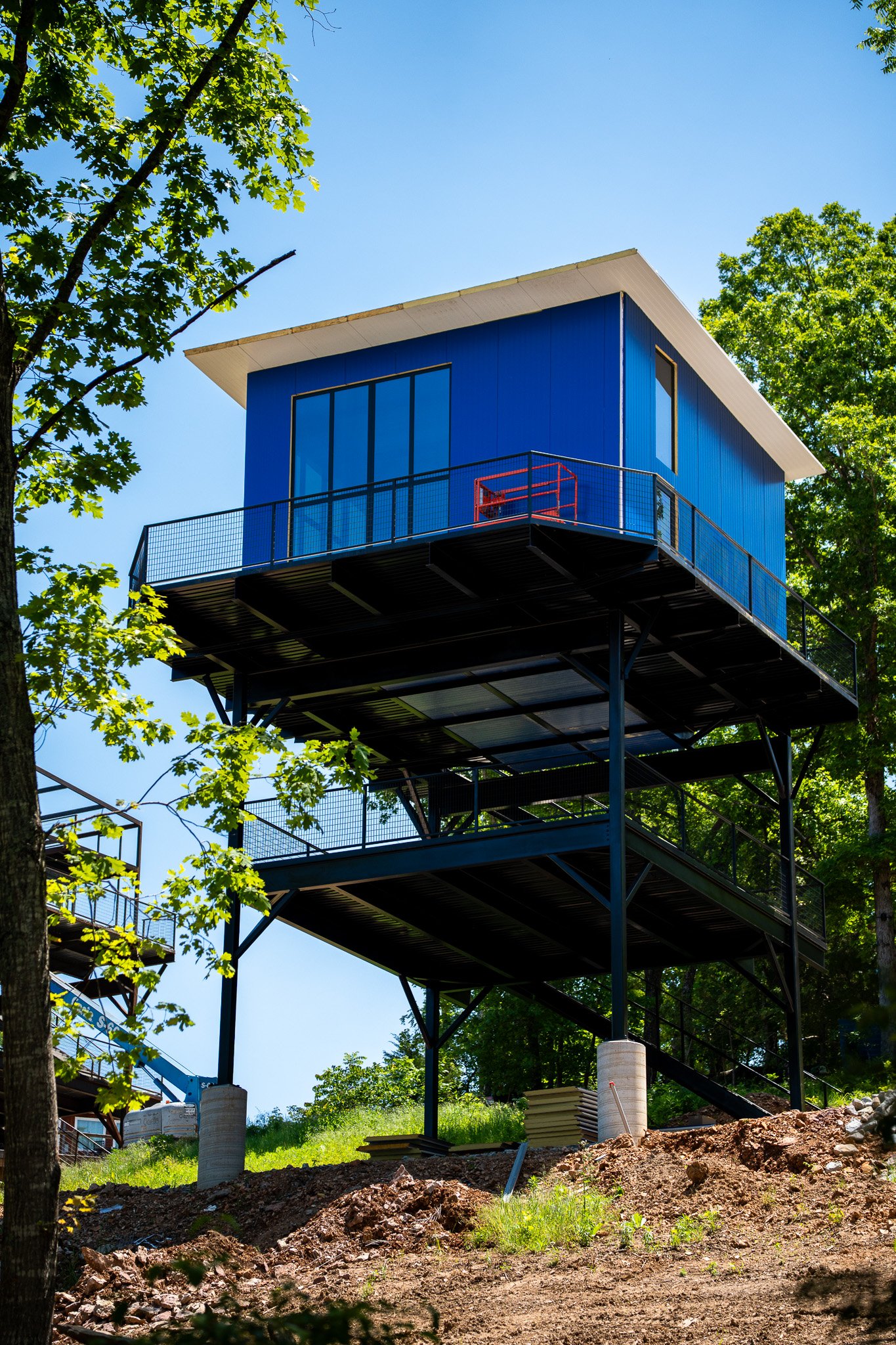 Construction site with a blue house-like structure elevated on stilts surrounded by green trees and a clear blue sky.