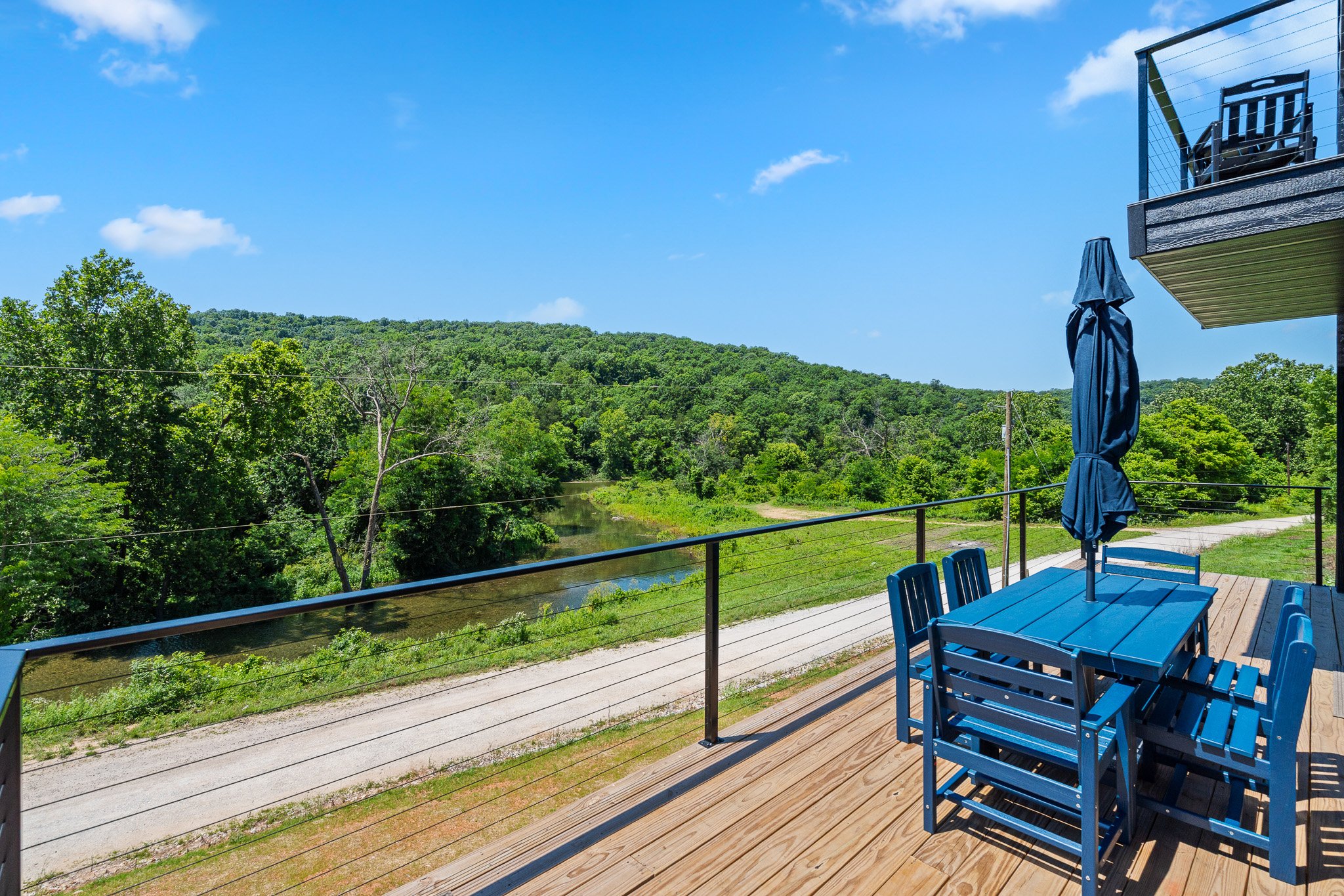 A balcony with a blue dining table, matching chairs, and an umbrella, overlooking a lush green landscape with trees and a river, under a blue sky with a few clouds.