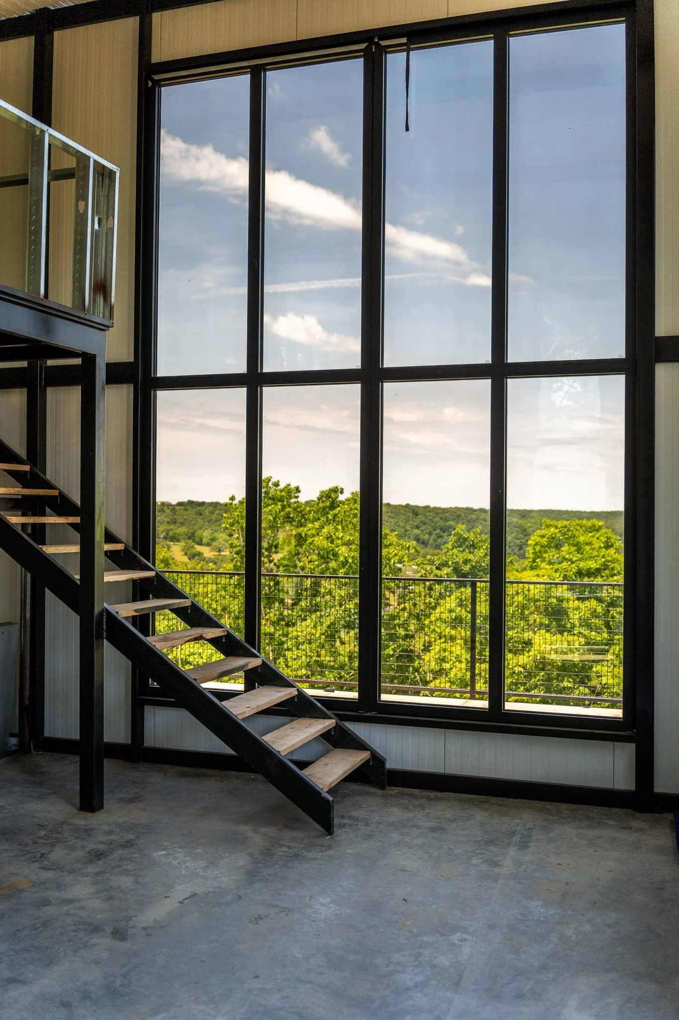 Interior of a building with a large floor-to-ceiling window showing a view of green trees and a cloudy sky. There are metal stairs with wooden steps on the left side of the image.