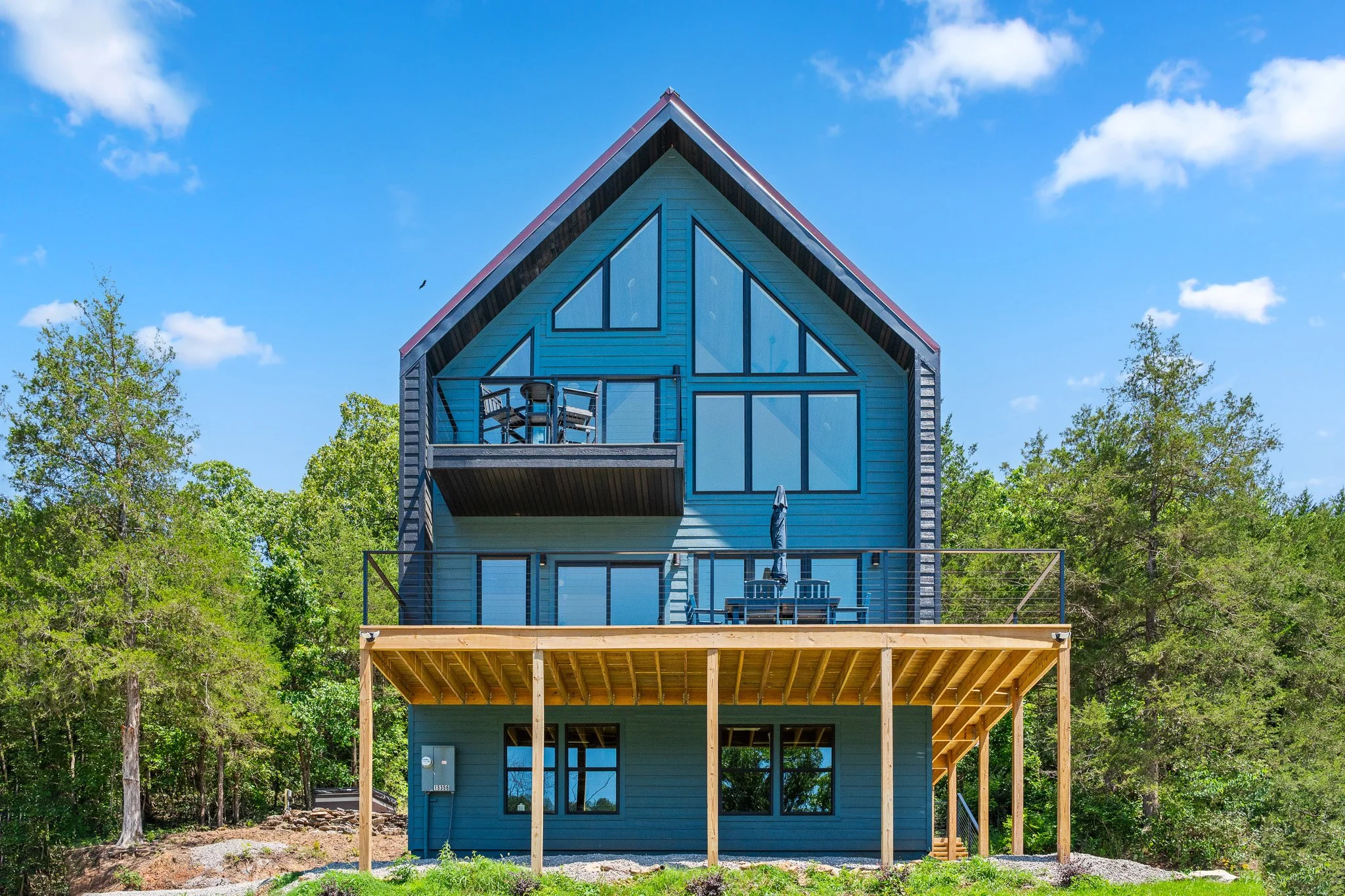 Front view of a modern blue house with large windows and a balcony, surrounded by green trees under a partly cloudy sky.