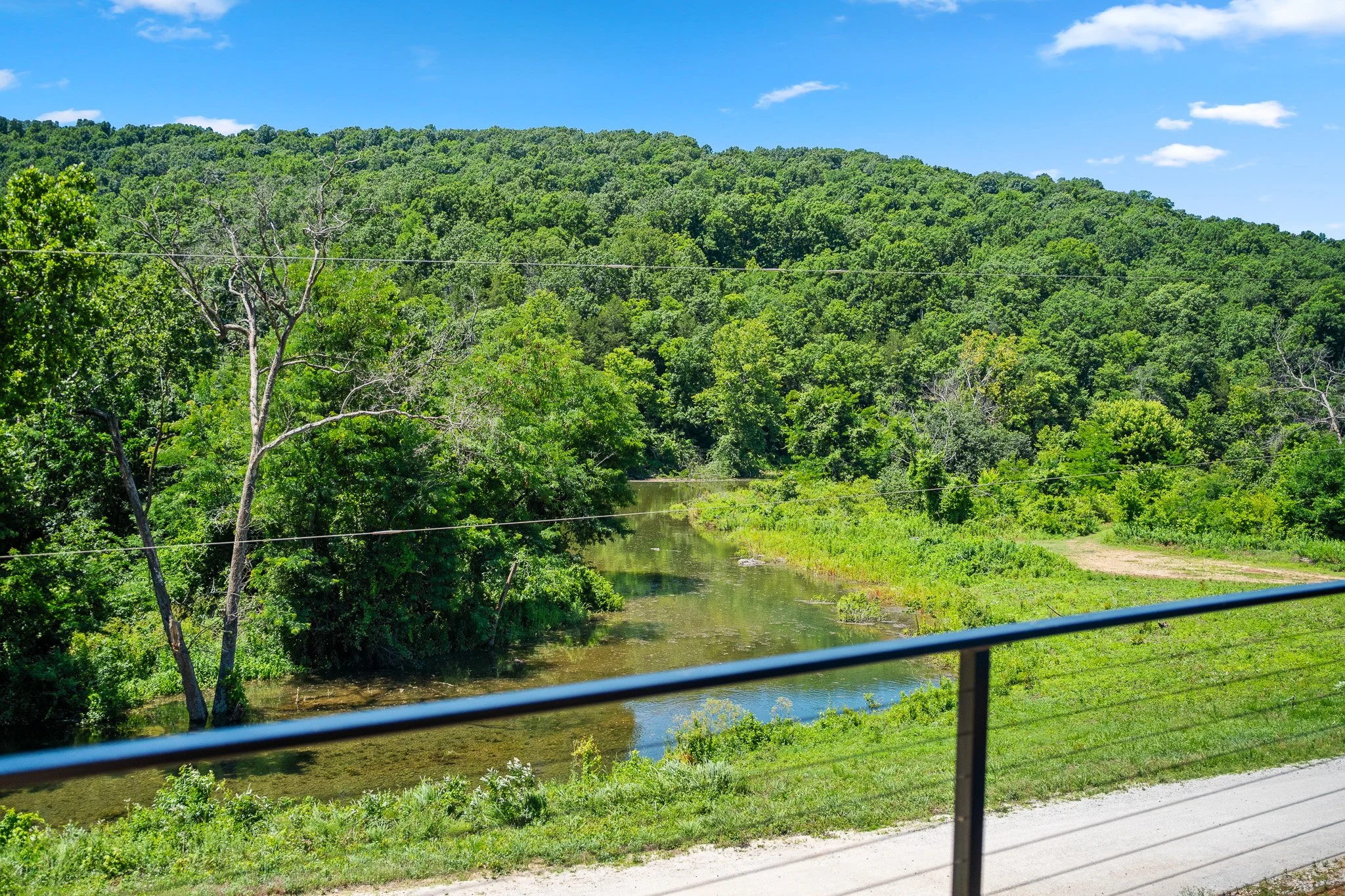 A scenic view of a lush green hill with trees, a river flowing at the base, and a blue sky with a few clouds, seen through a vehicle's window with a black railing in the foreground.
