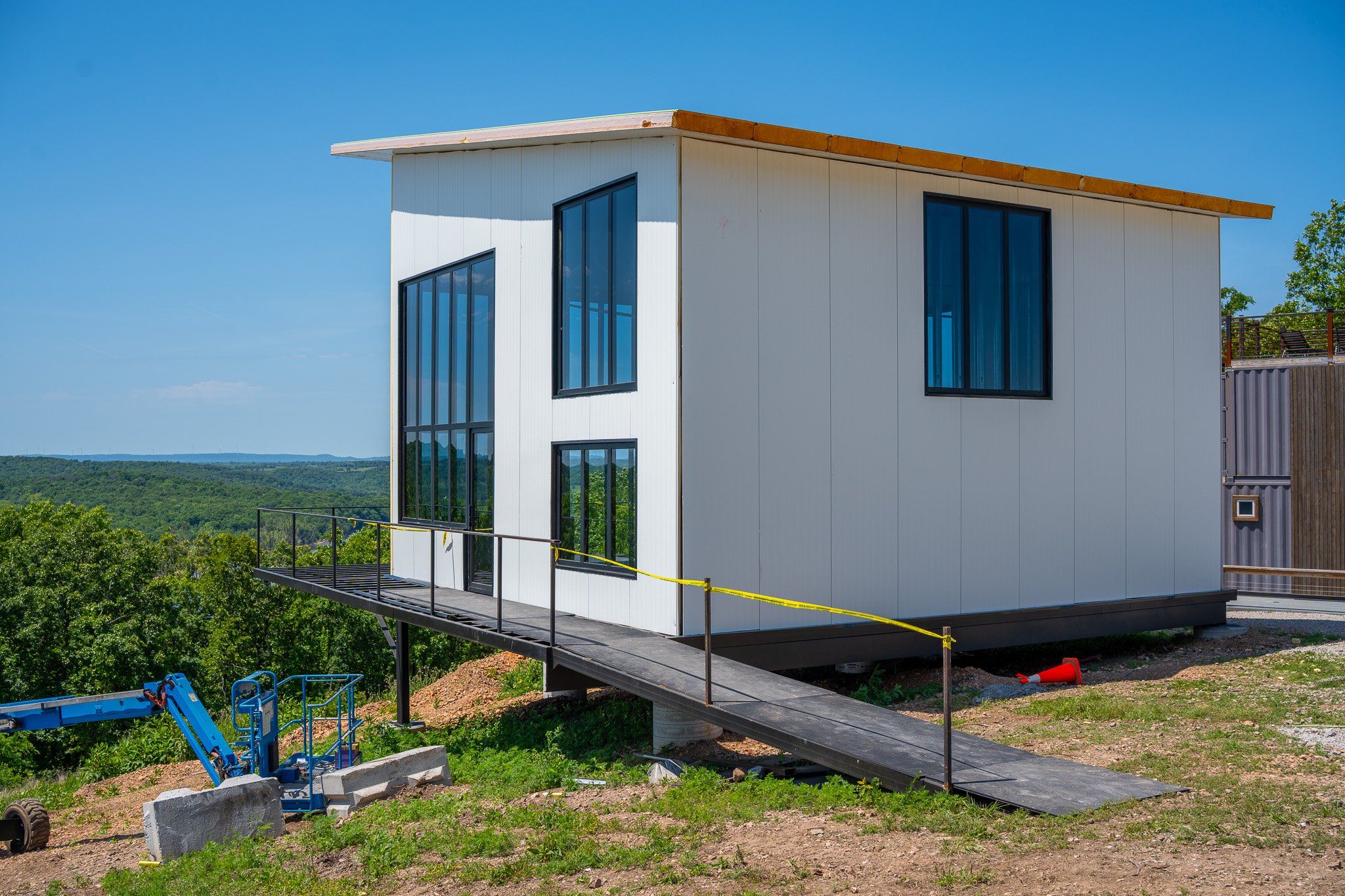 Modern house under construction with large windows and a slanted roof, surrounded by trees and open landscape, with construction equipment nearby.