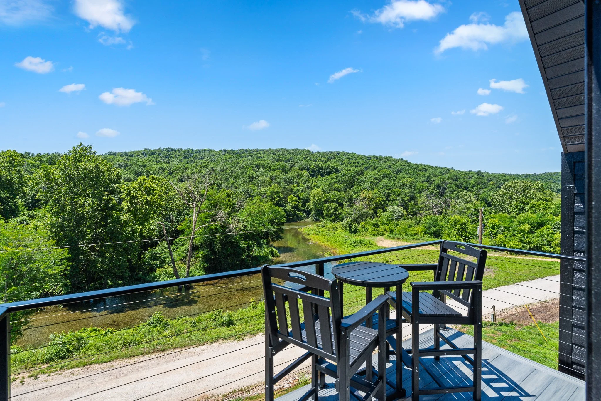 View from a balcony with black metal chairs and table overlooking a lush green landscape with a river and trees under a blue sky with clouds.