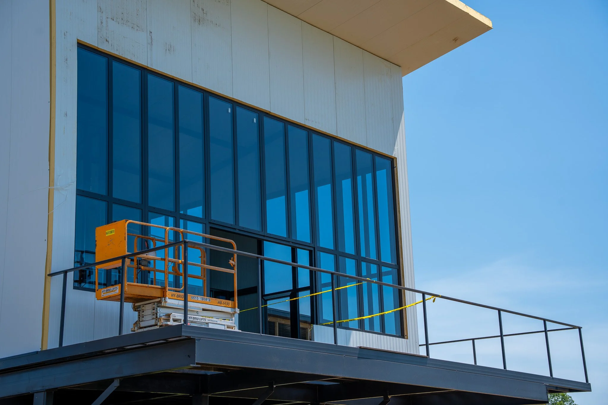 A building under construction with glass windows and a construction lift on a balcony, with a blue sky in the background.
