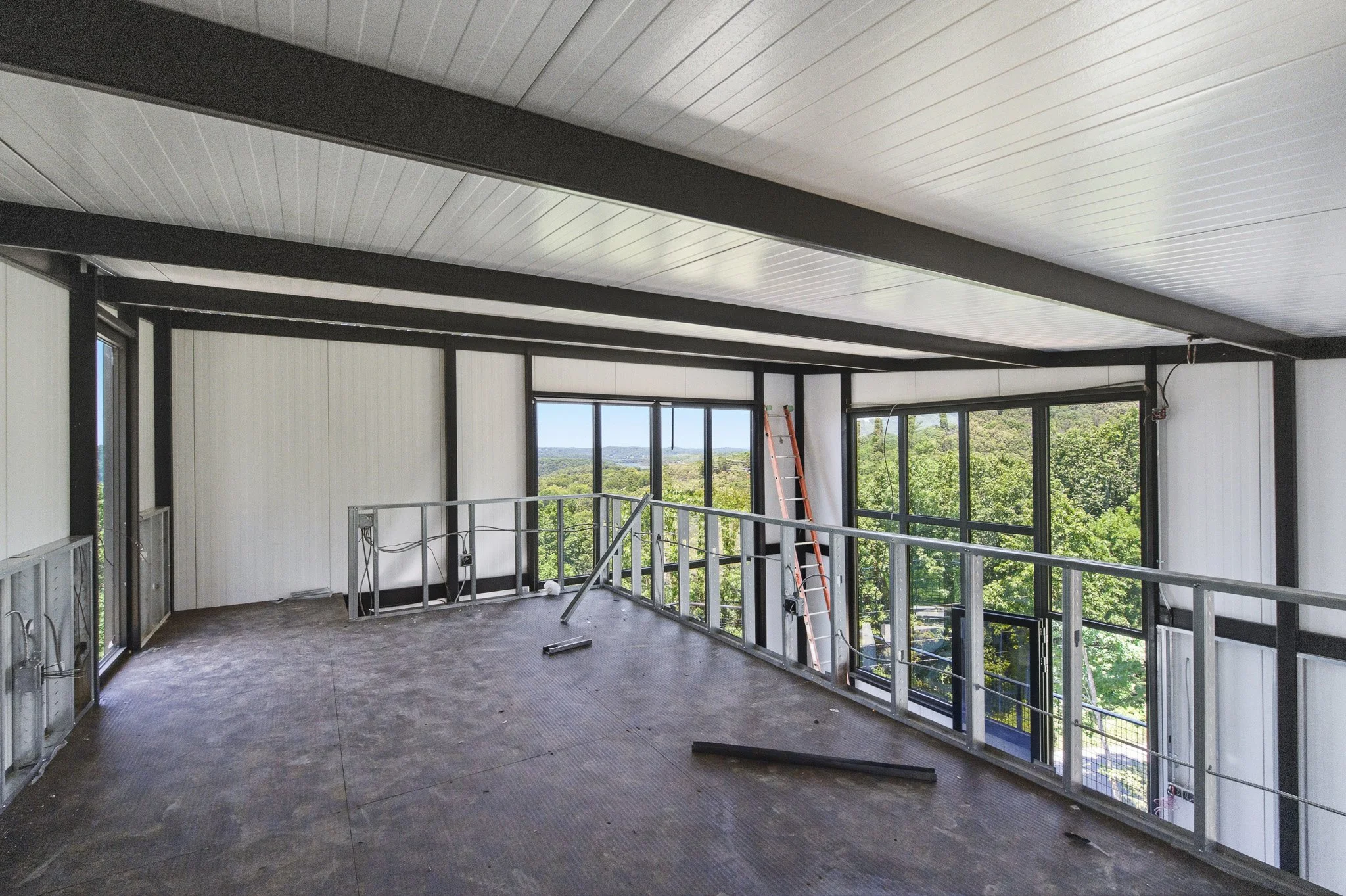 Interior view of a building under construction with large windows, metal framing, a ladder, and a view of trees and landscape outside.