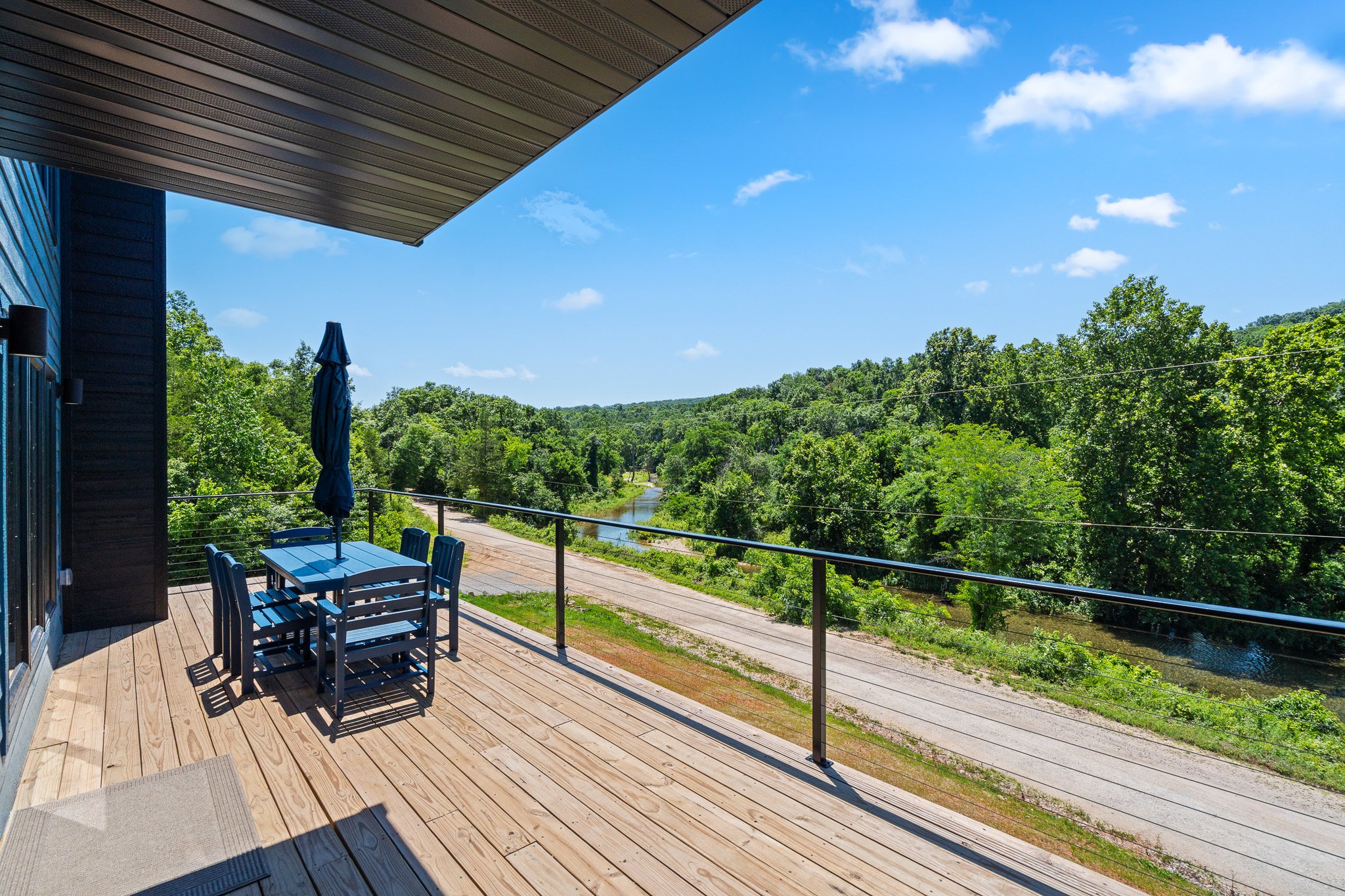 A wooden deck with outdoor tables and chairs, overlooking lush green trees and a river under a partly cloudy blue sky.