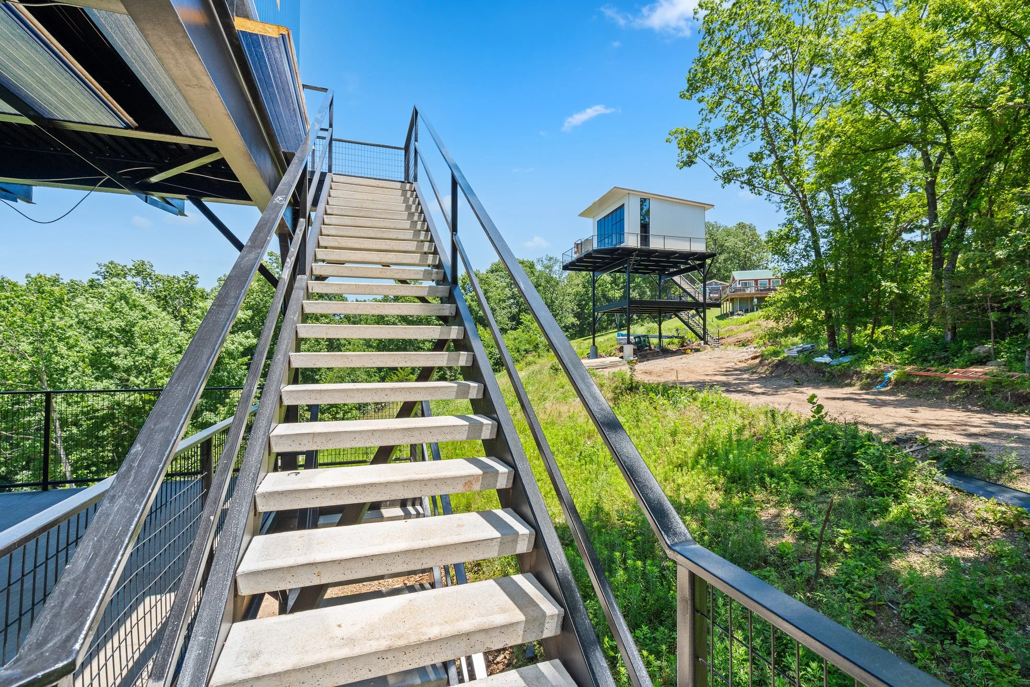 Outdoor view of a stairway leading up to a modern tiny house built on stilts among green trees, with a dirt pathway, under a blue sky with some clouds.