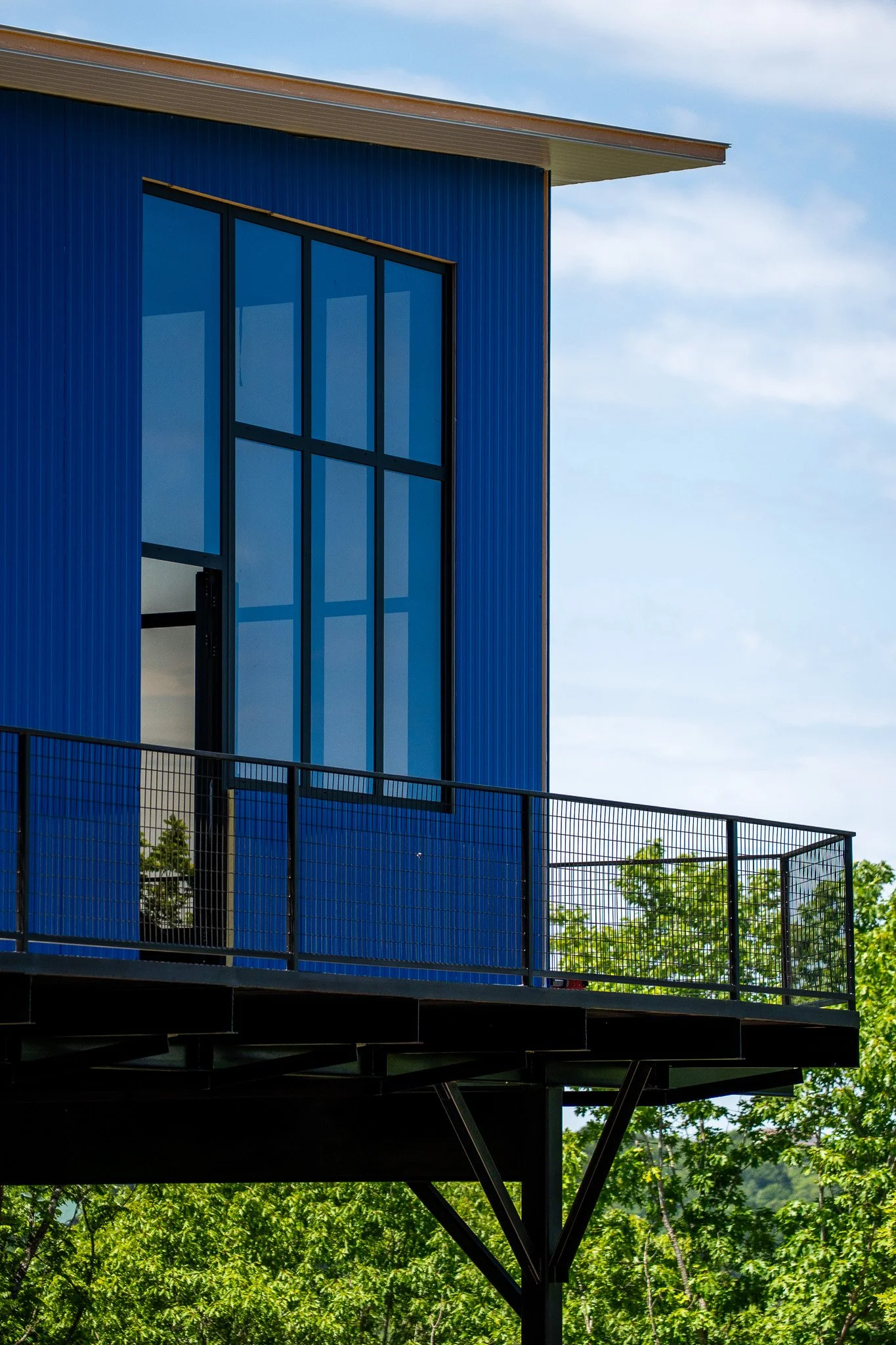 A modern building with blue exterior walls and large glass windows, situated above a lush green forest, with a metal fence on the balcony railing.