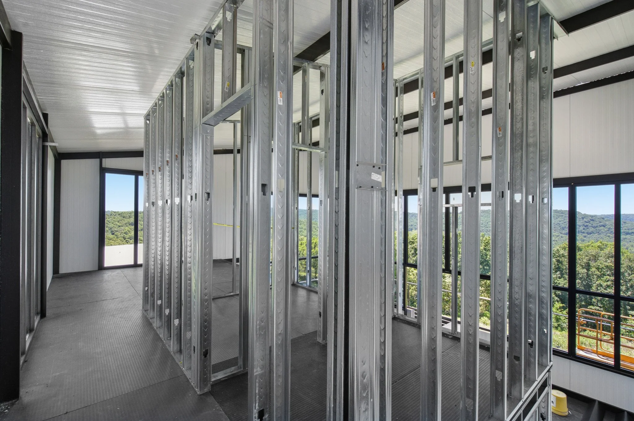 Interior of a building under construction, showing metal wall framing with large windows and a view of greenery outside.