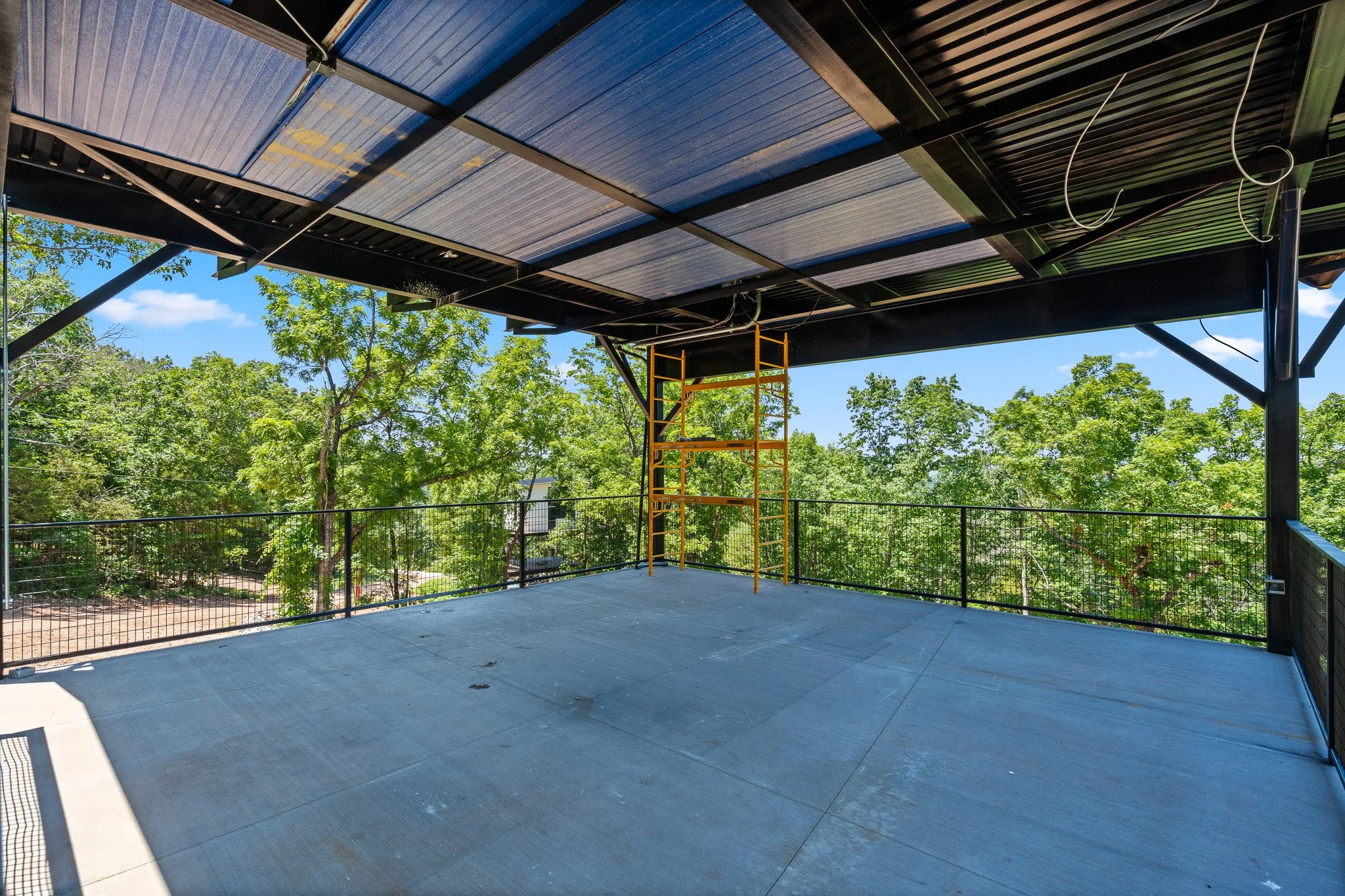 Empty outdoor balcony or rooftop with a black metal railing, a yellow scaffold, and a blue sky with trees in the background.