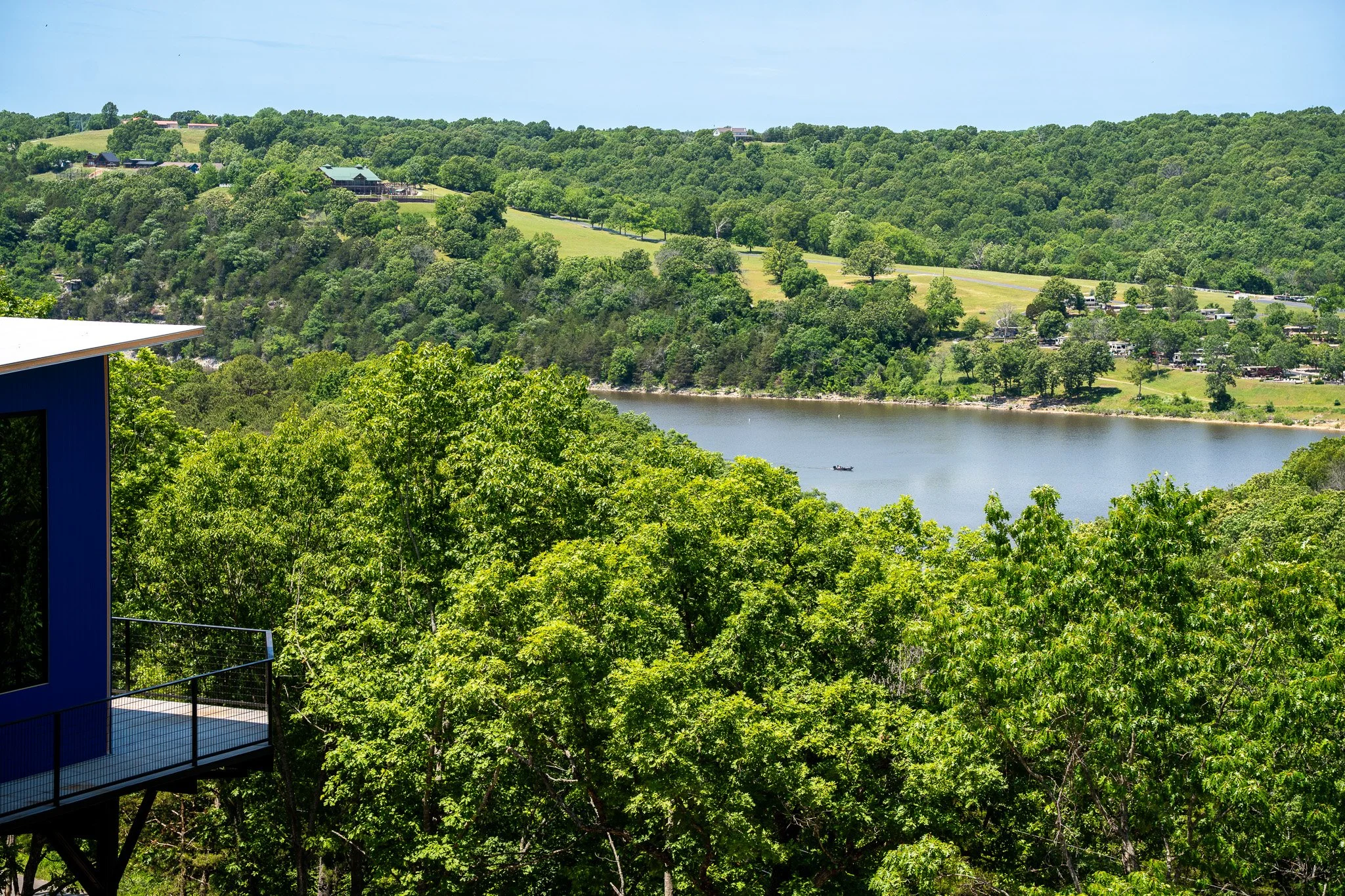 Scenic view of a green hillside with trees, houses, and a body of water in the foreground. A small boat is on the water, and there is a building with a balcony in the lower left corner.
