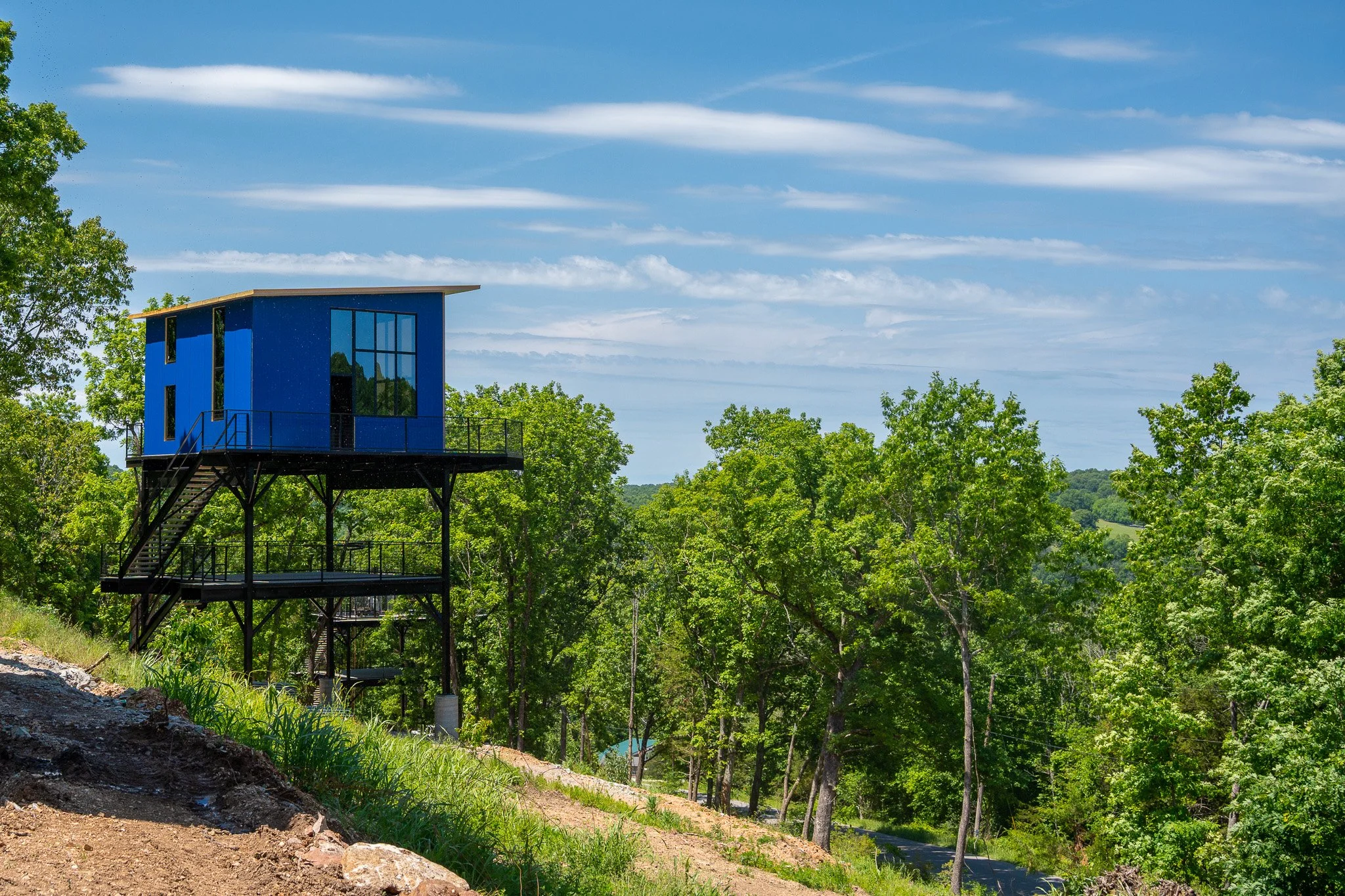A modern blue house elevated on stilts surrounded by green trees under a blue sky with wispy white clouds.