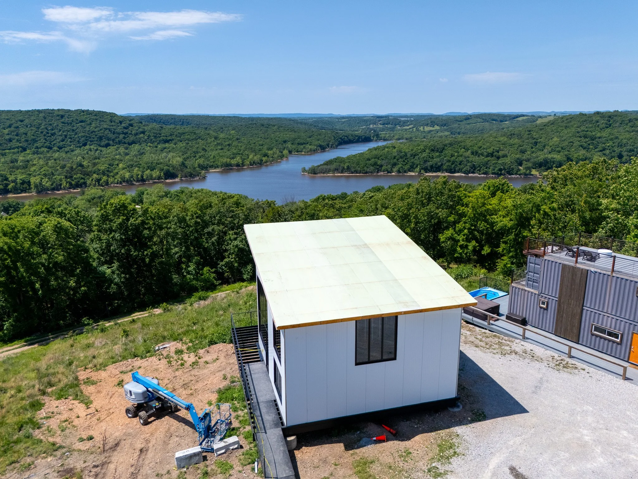 A modern white house under construction near a river with green forested hills in the background, construction equipment, and a paved driveway.