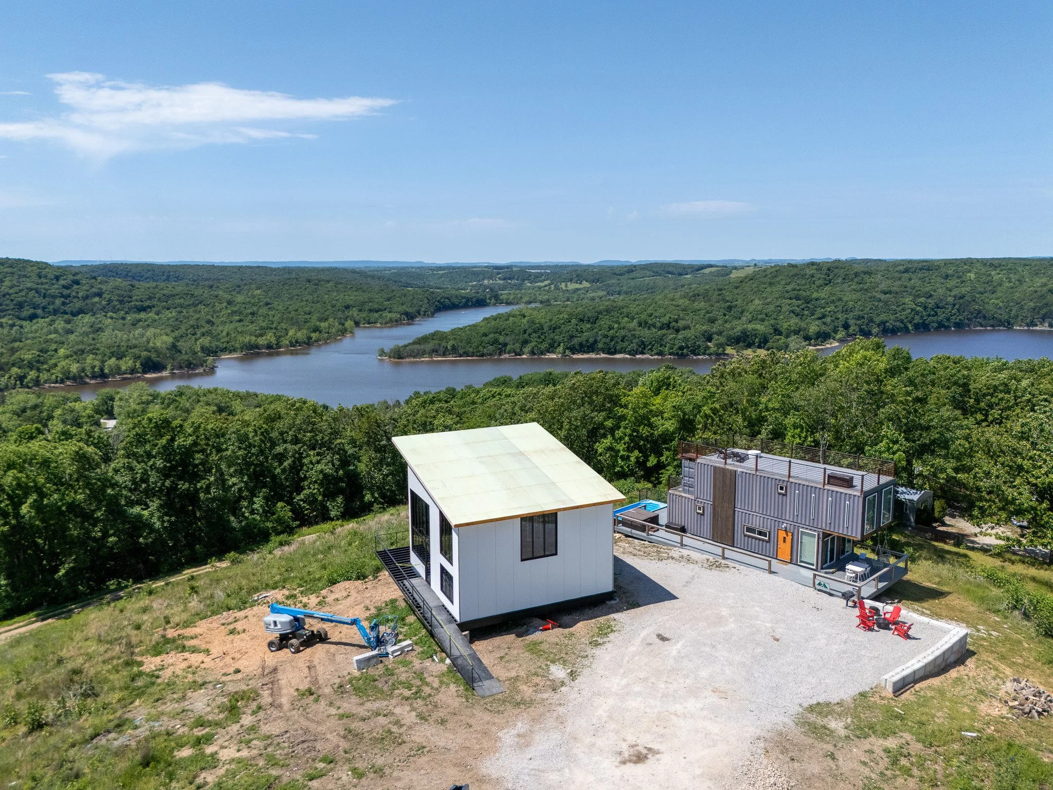 Aerial view of a construction site with partially built modern small house, construction equipment, and a background of a river and green forested rolling hills under blue sky.