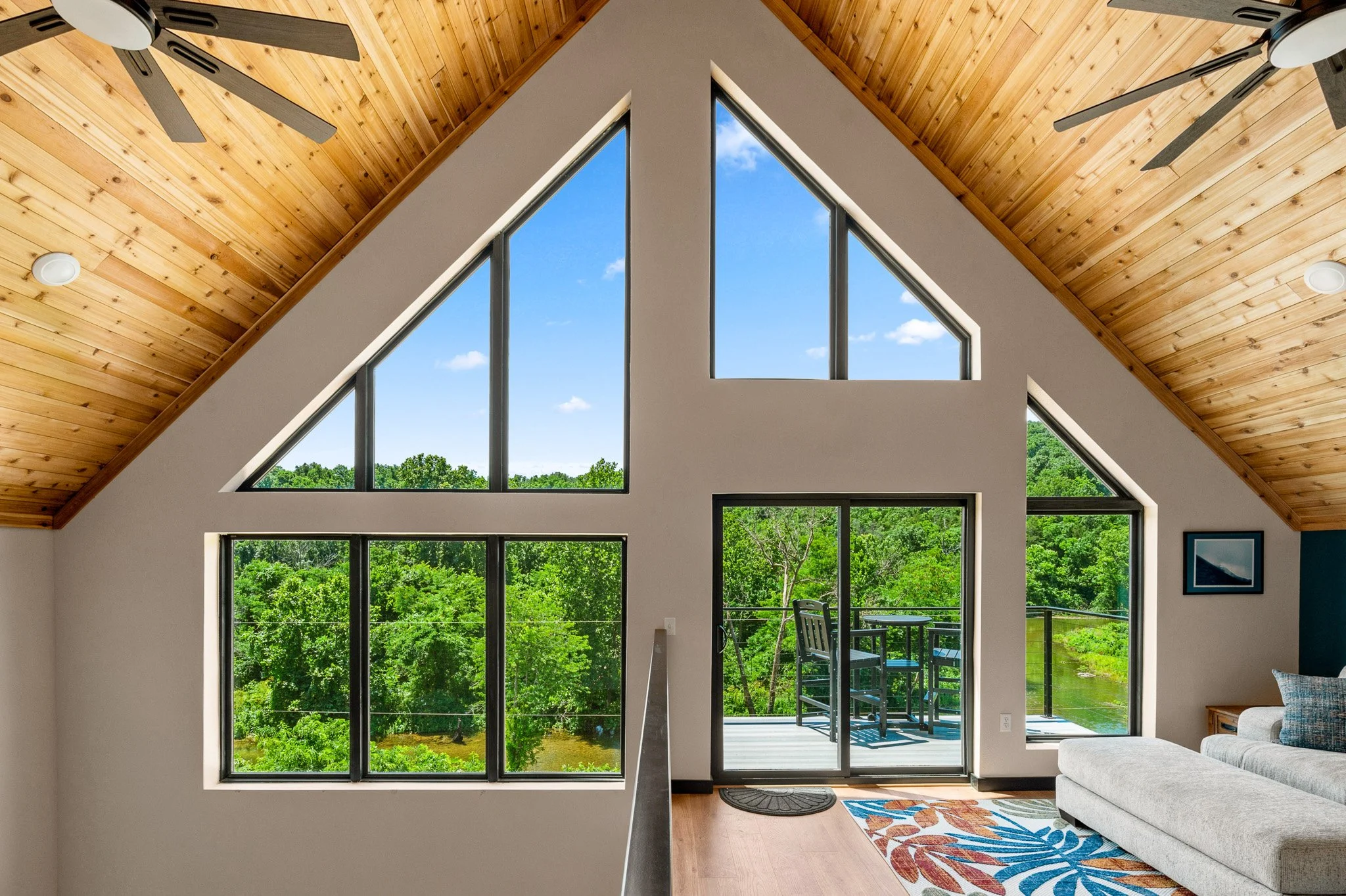 Living room with large triangular windows, wooden vaulted ceiling, ceiling fans, and a view of green trees outside.