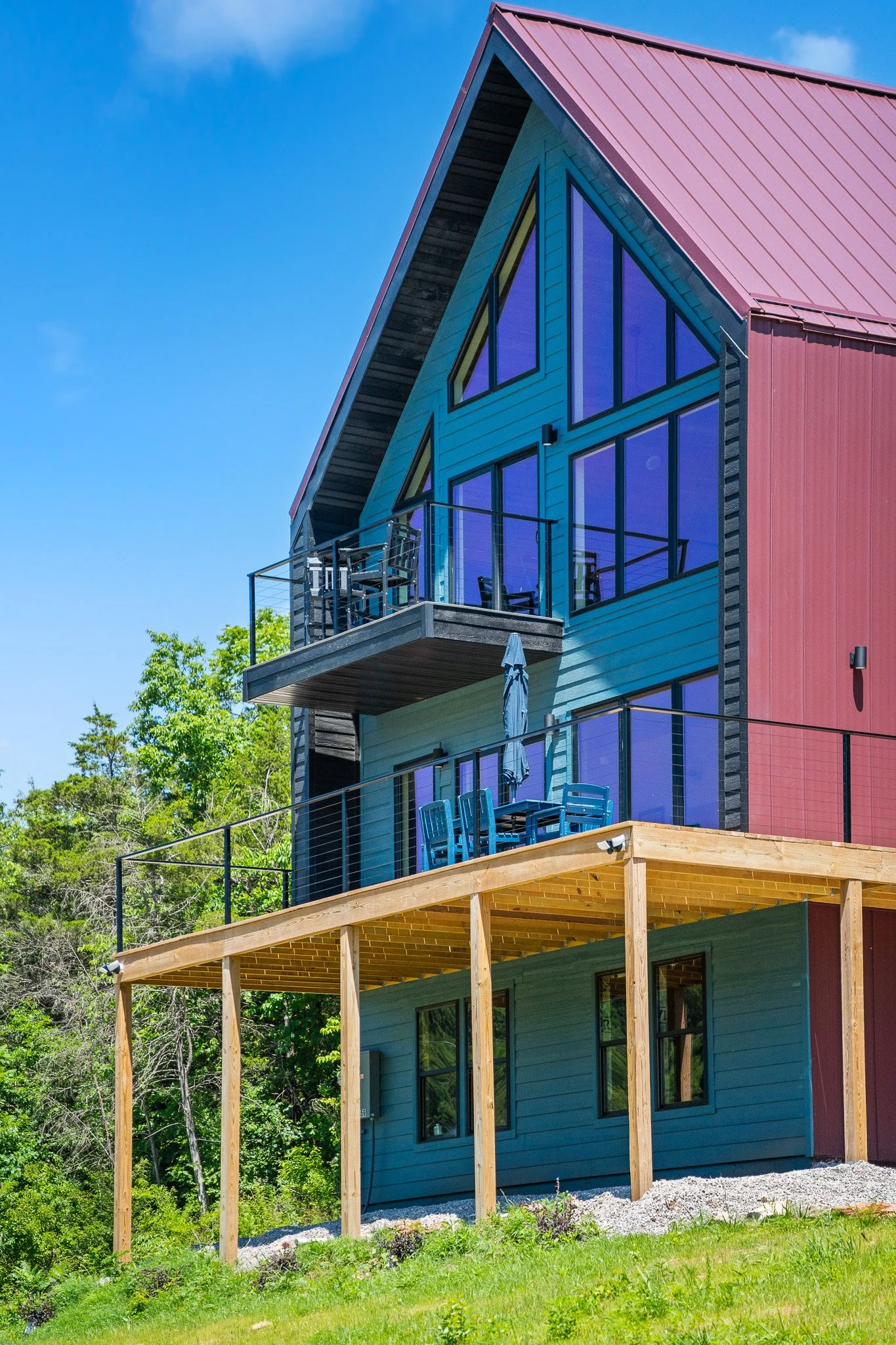 The image shows a colorful, modern house with large triangular windows and a balcony on a bright, sunny day. The house is painted in shades of blue and red, with black framing around the windows. There is a wooden deck below the house with some outdo