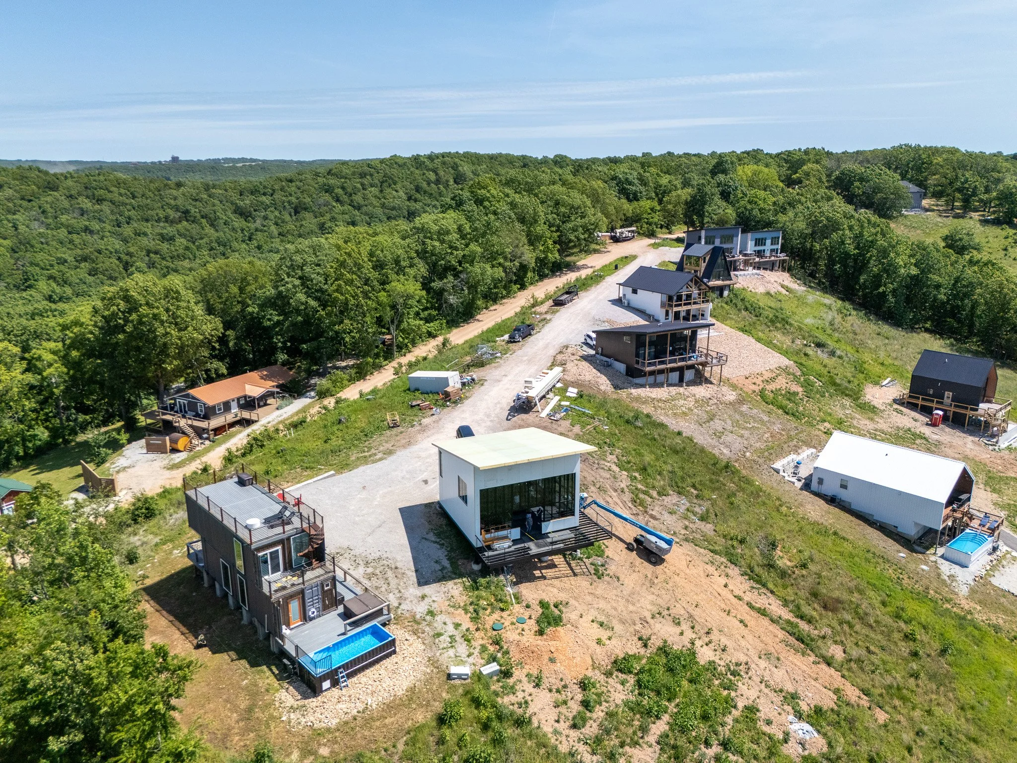 Aerial view of a residential area with modern houses, some with decks and swimming pools, surrounded by lush green trees and dirt roads on a clear day.