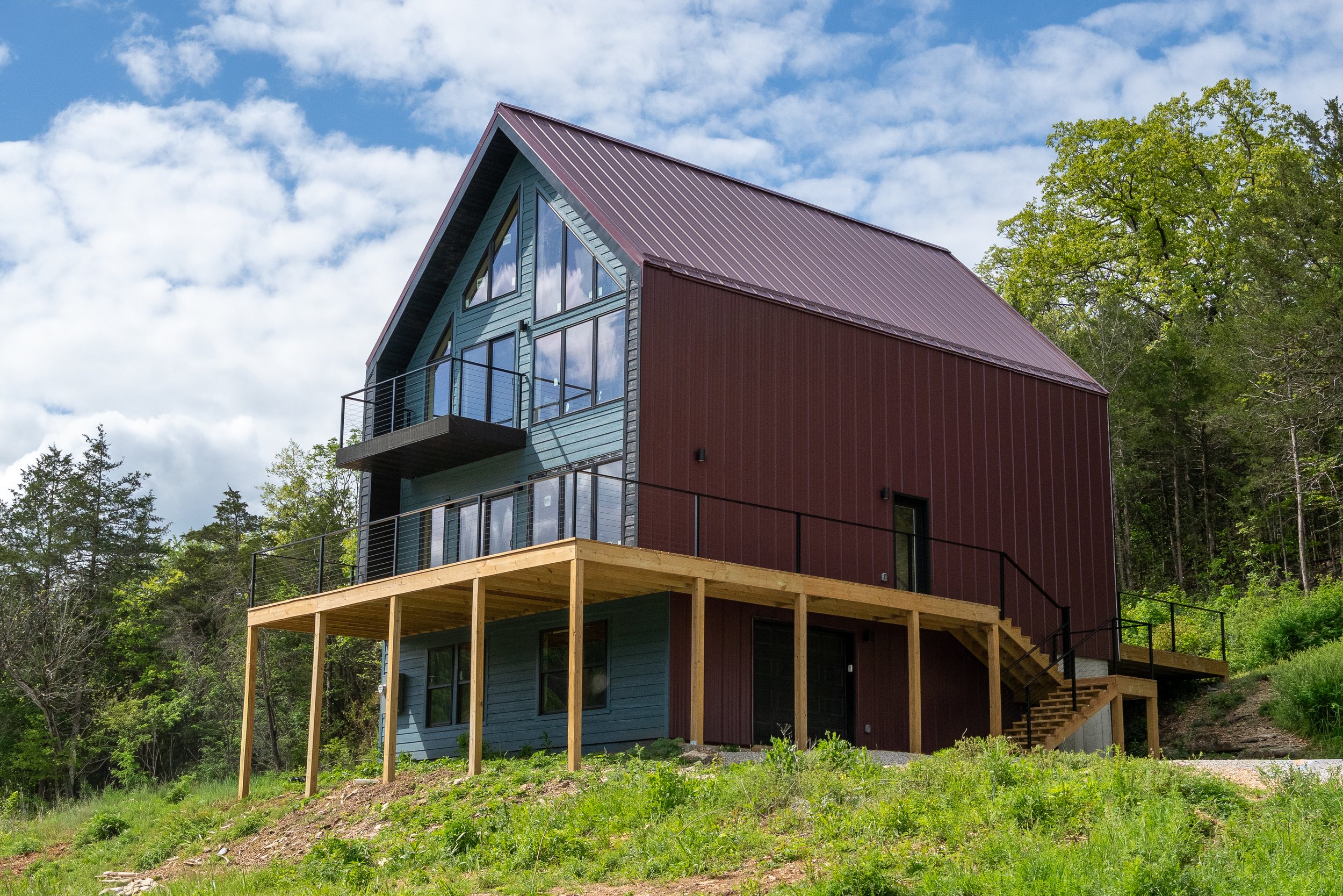 A modern house with large glass windows and a steep roof, built on a hillside with a wooden deck and stairs, surrounded by green trees and a partly cloudy sky.