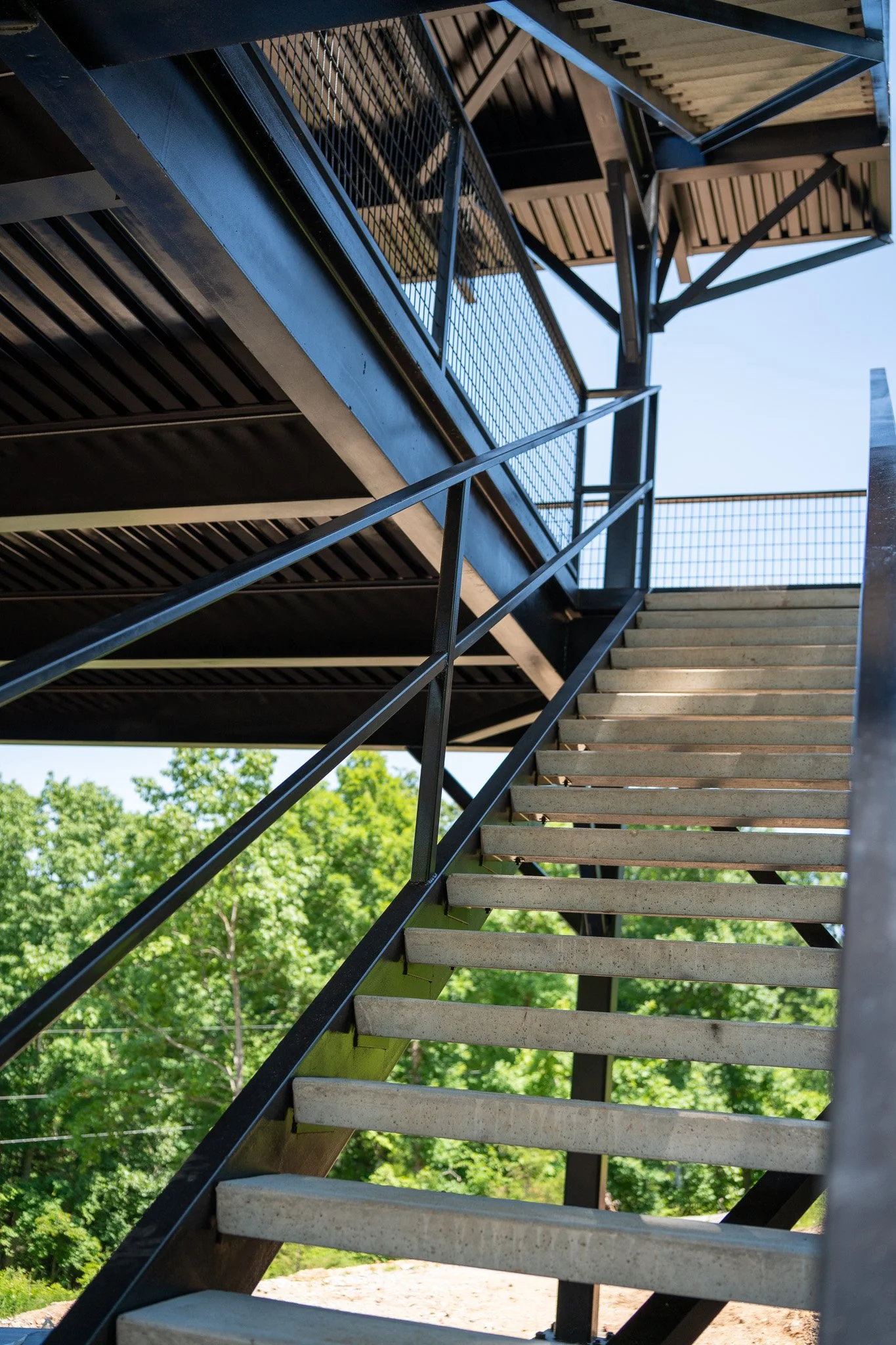 Outdoor metal staircase with wooden steps against a backdrop of green trees and a clear blue sky.