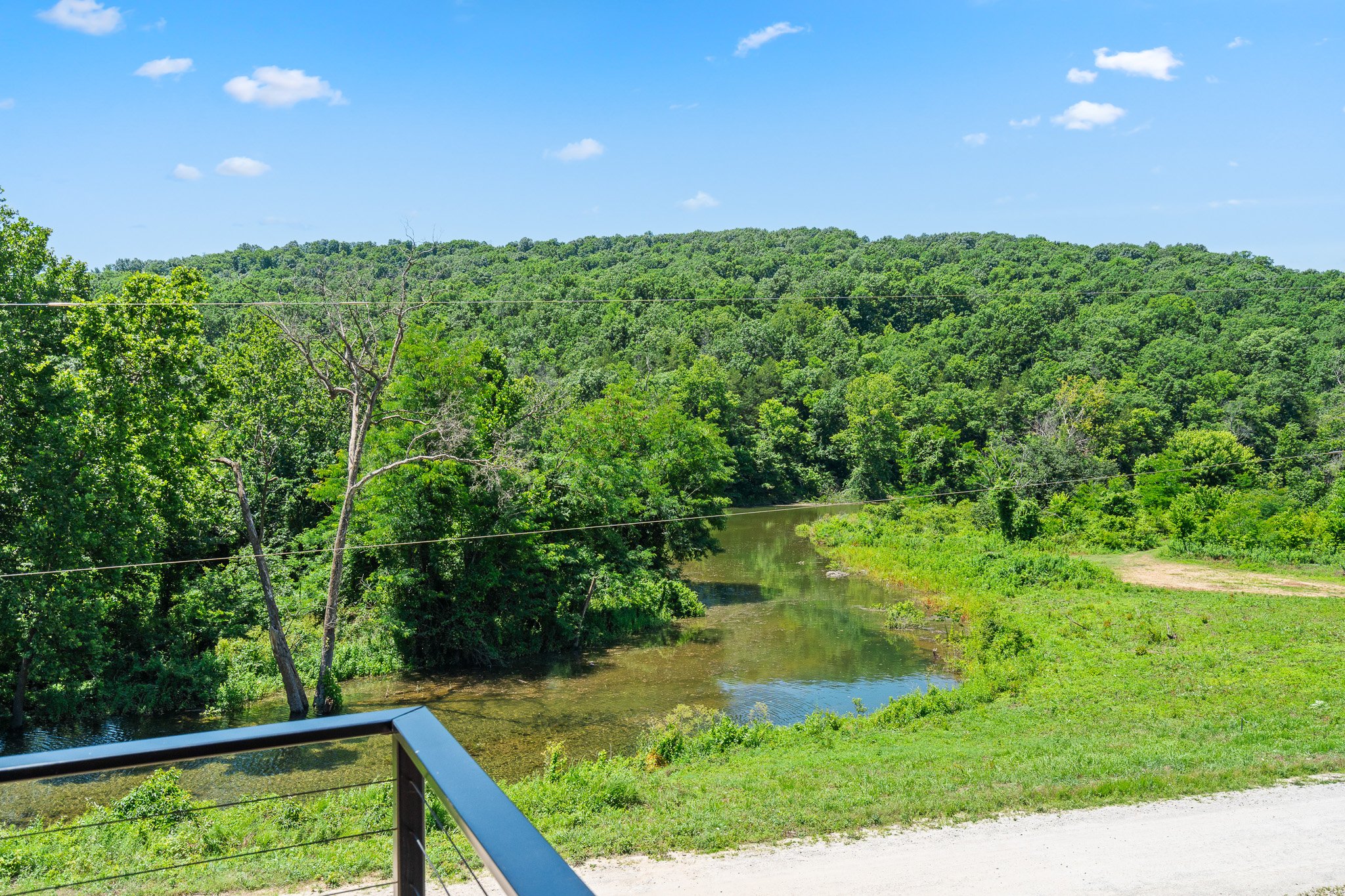 A scenic view of a river running through a lush green forest under a bright blue sky with scattered white clouds.