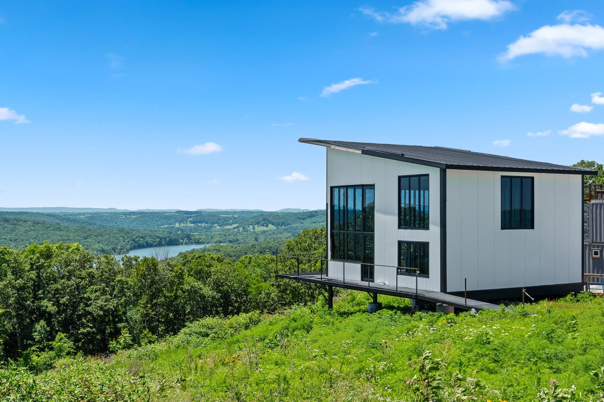 Modern house with large windows on a hillside overlooking a river and green landscape under a blue sky with scattered clouds.