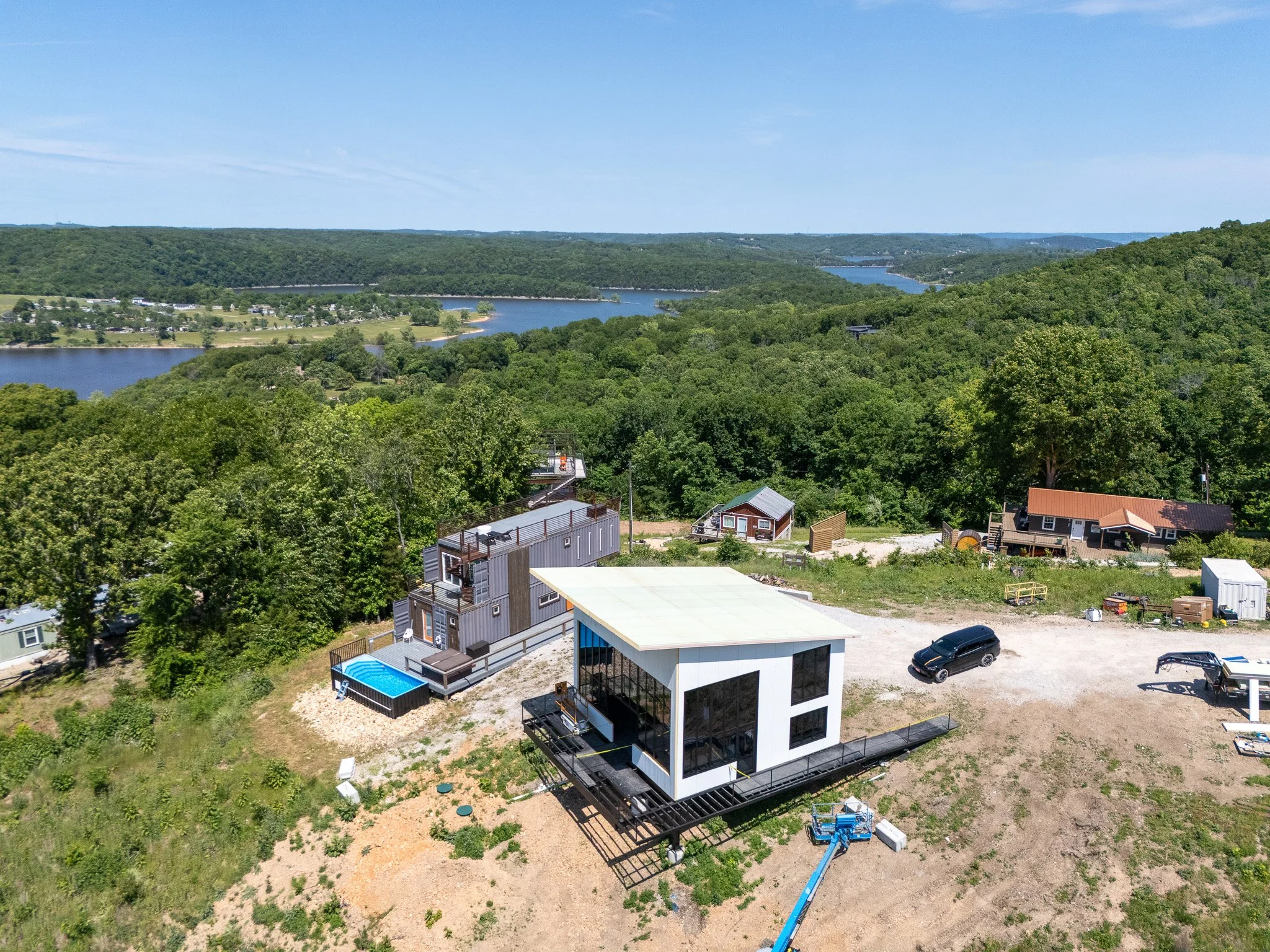 A residential area with a modern, multi-story house featuring large glass windows, a deck, and a swimming pool, set amidst green trees, with a lake and rolling hills in the background under a clear blue sky.