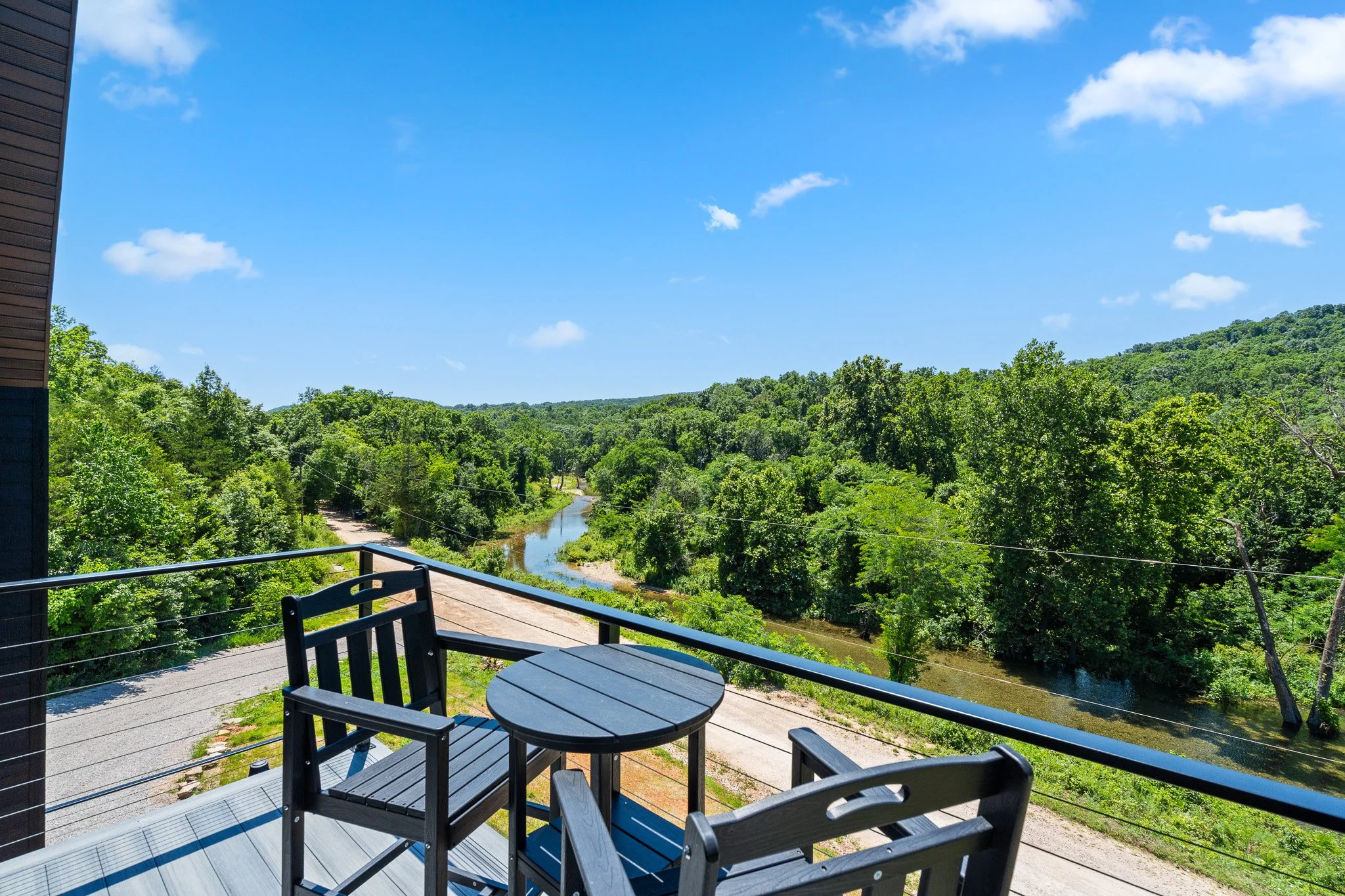 Balcony with a round black table and chairs overlooking a lush green landscape with a river and hills under a blue sky with a few white clouds.