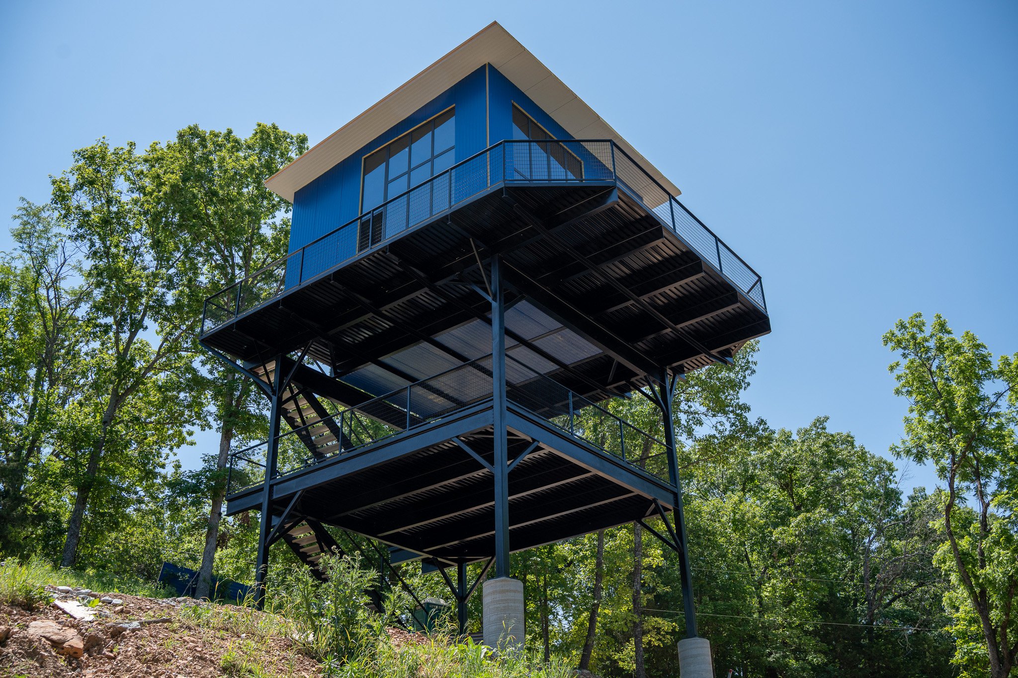 A modern blue treehouse built on a metal structure with multiple decks, situated among green trees under a clear blue sky.