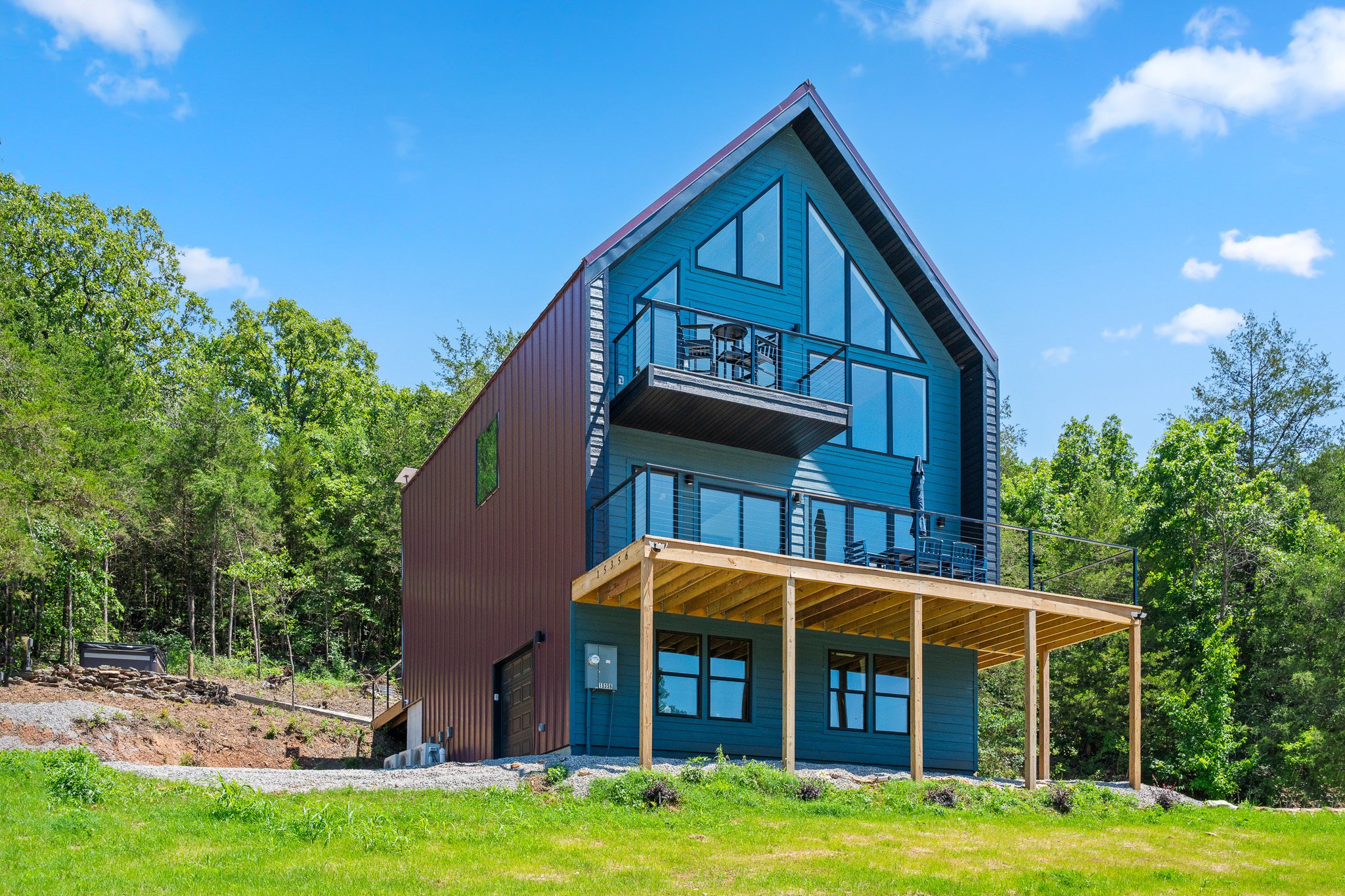 A modern house with blue siding and large glass windows, situated on a grassy area with trees in the background, under a bright blue sky with scattered clouds.