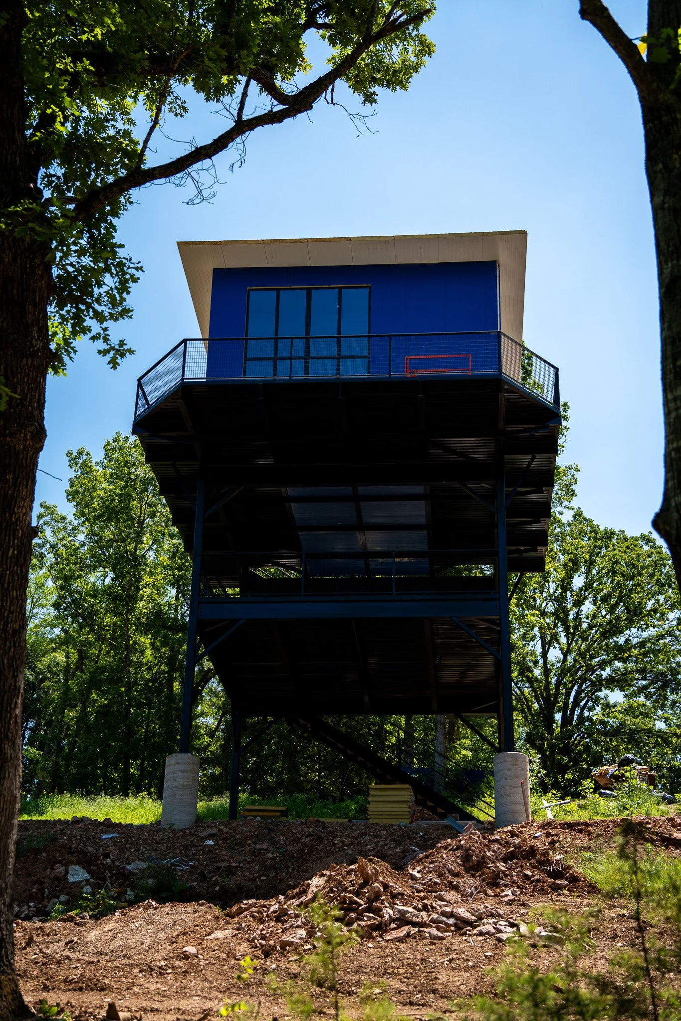 A modern blue building with glass doors and a wraparound balcony, elevated on metal supports, surrounded by trees and a clear blue sky.
