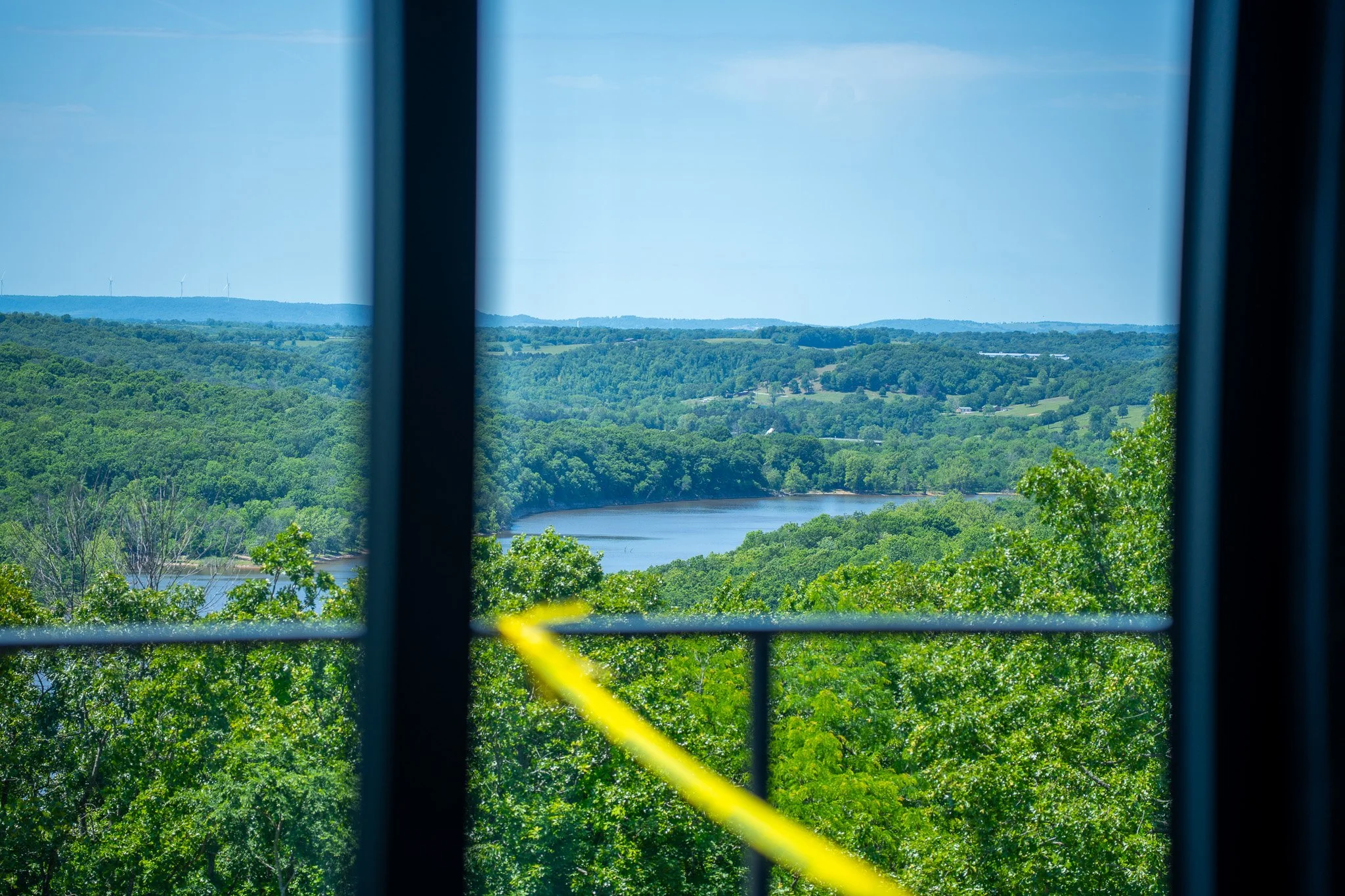 View of lush green trees, a body of water, and rolling hills with wind turbines seen through a window with a yellow safety line.