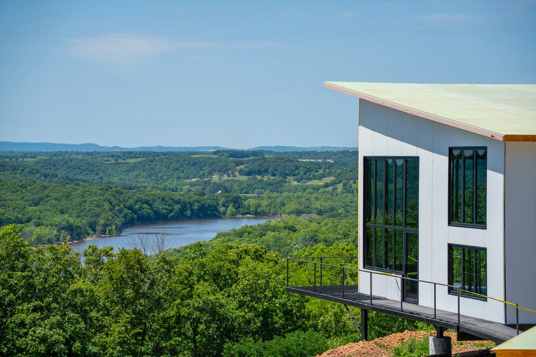 Modern house with large windows and a balcony overlooking a lush green landscape and a river, under a blue sky.